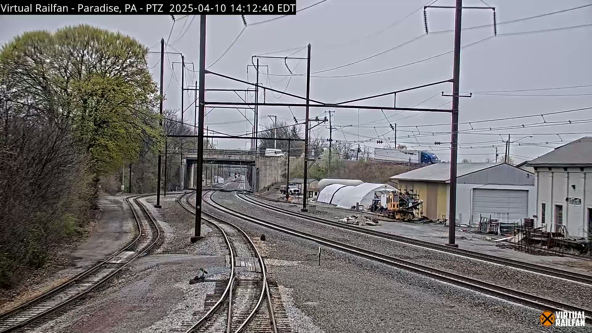 Several sets of train tracks stretch into the distance under an overcast sky, passing near a road, buildings, and some trees showing new spring growth.