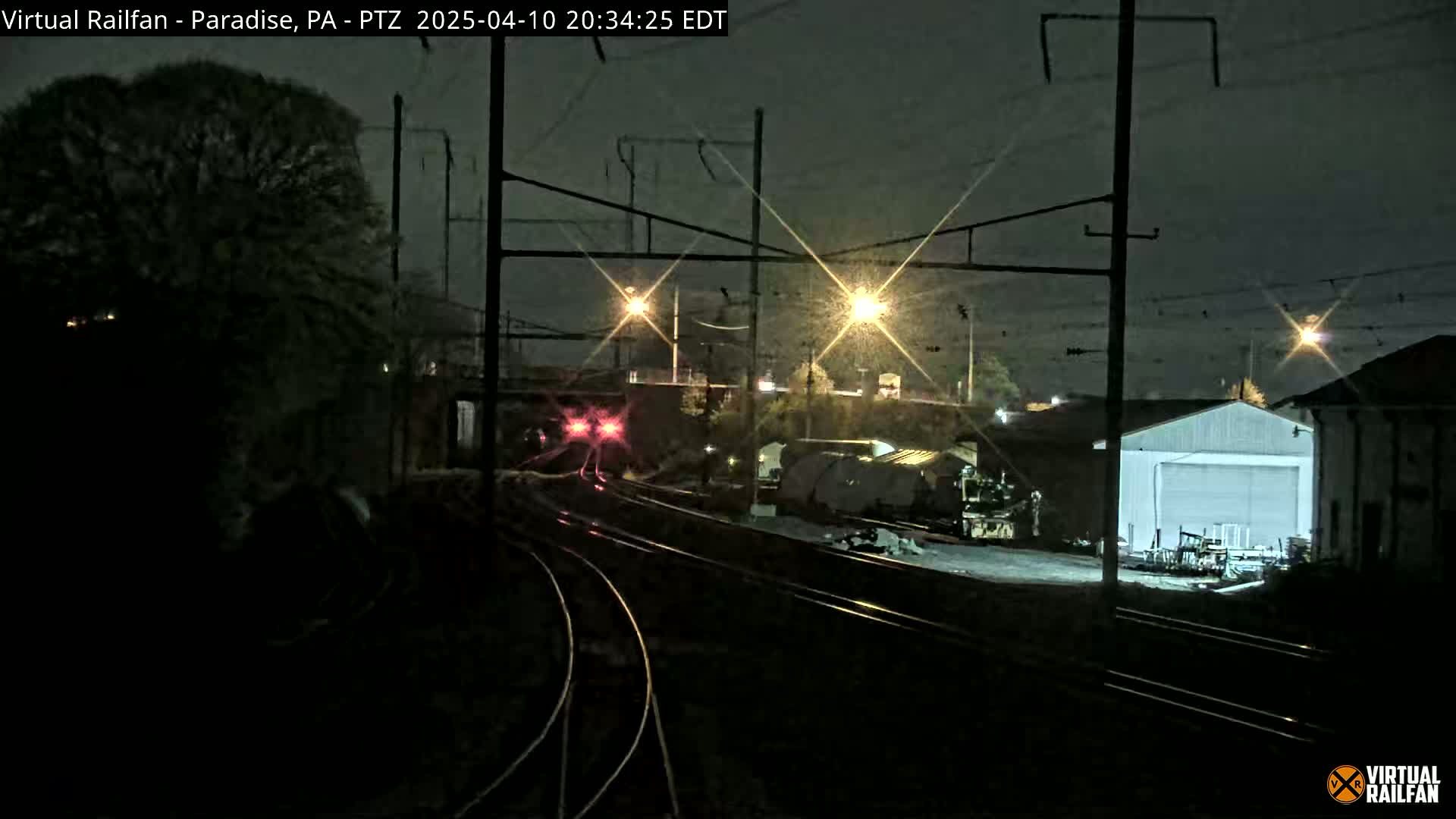 A nighttime view of railroad tracks and railway infrastructure under overcast skies, showing a train in the distance and a building nearby.