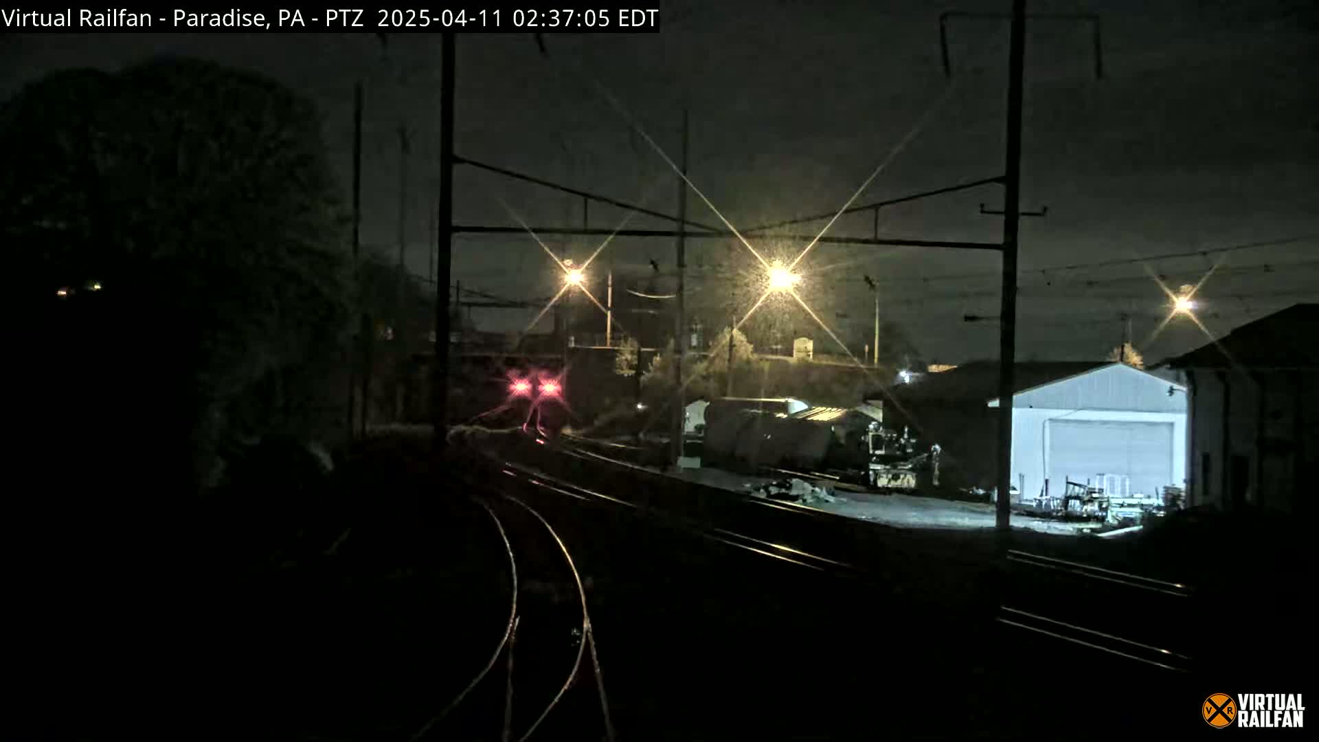 A nighttime view of a train yard with railway tracks, a train in the distance, and a brightly lit building under a dark sky.