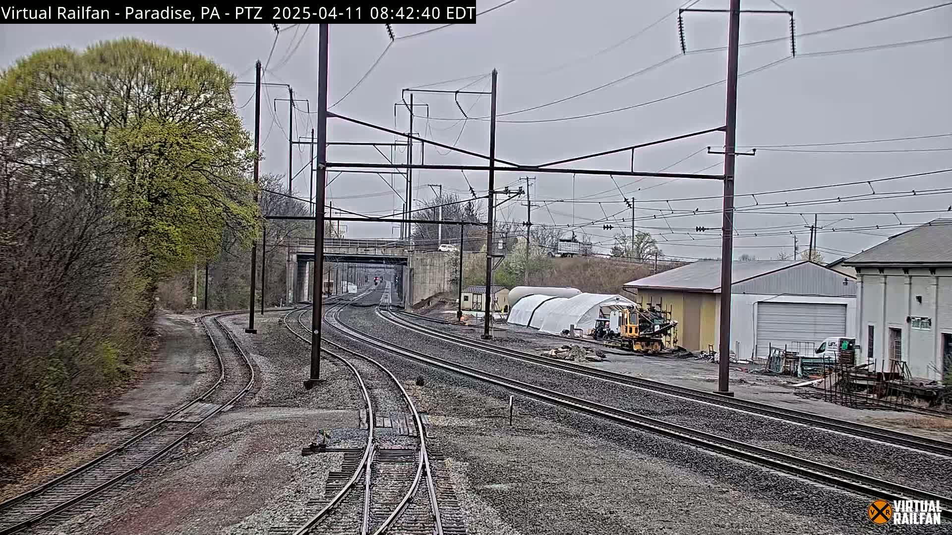 Multiple sets of train tracks extend into the distance under an overcast sky, passing near buildings and some construction equipment, with trees budding leaves in the background.