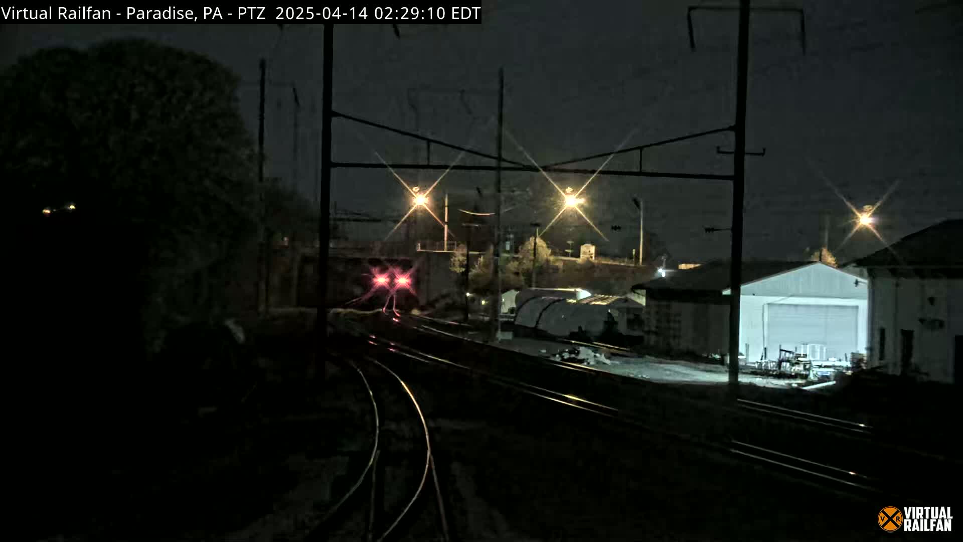A train with red lights is passing through a rail yard at night.