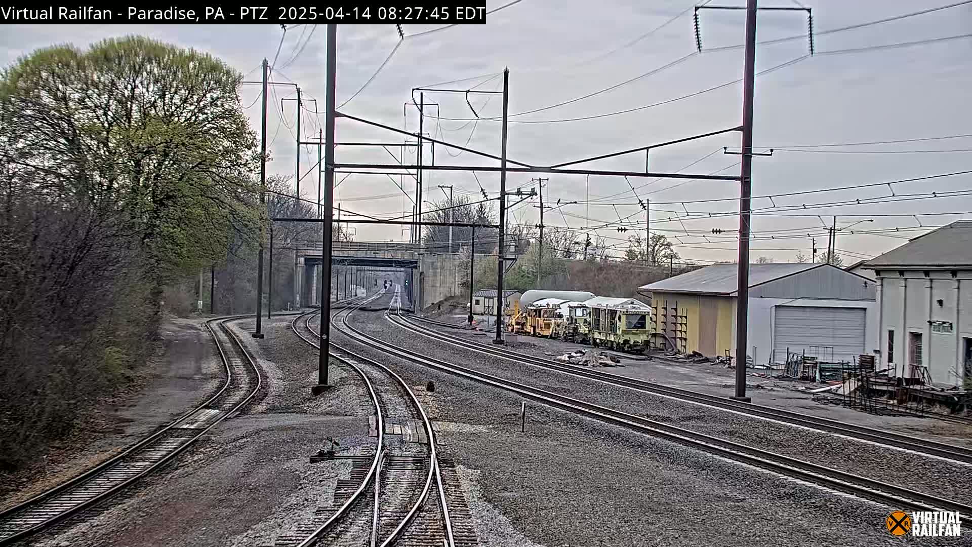 Multiple sets of train tracks curve through a landscape of trees and industrial buildings under an overcast sky.