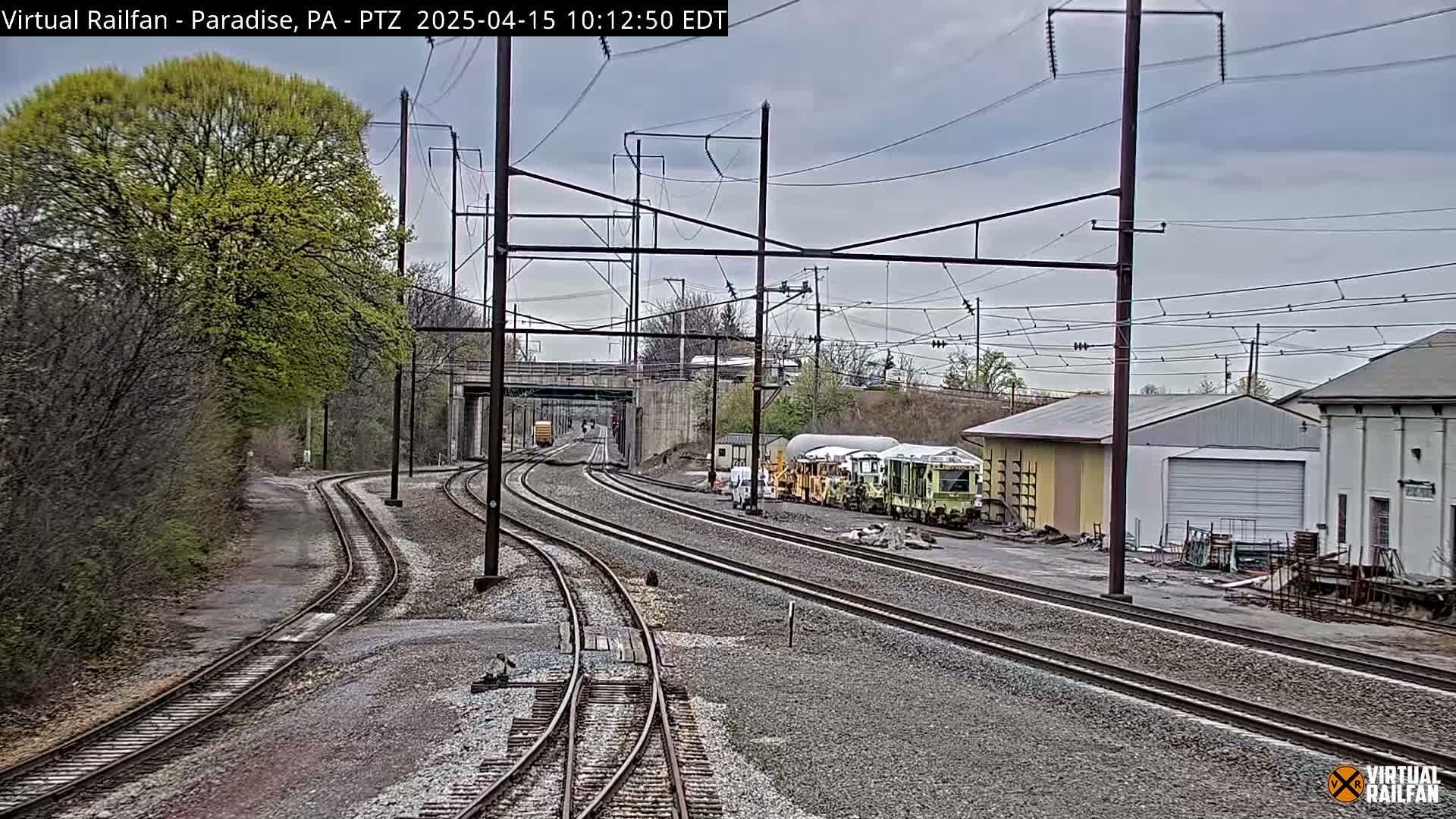 Several sets of train tracks converge near a rail yard with buildings and overgrown vegetation under an overcast sky.