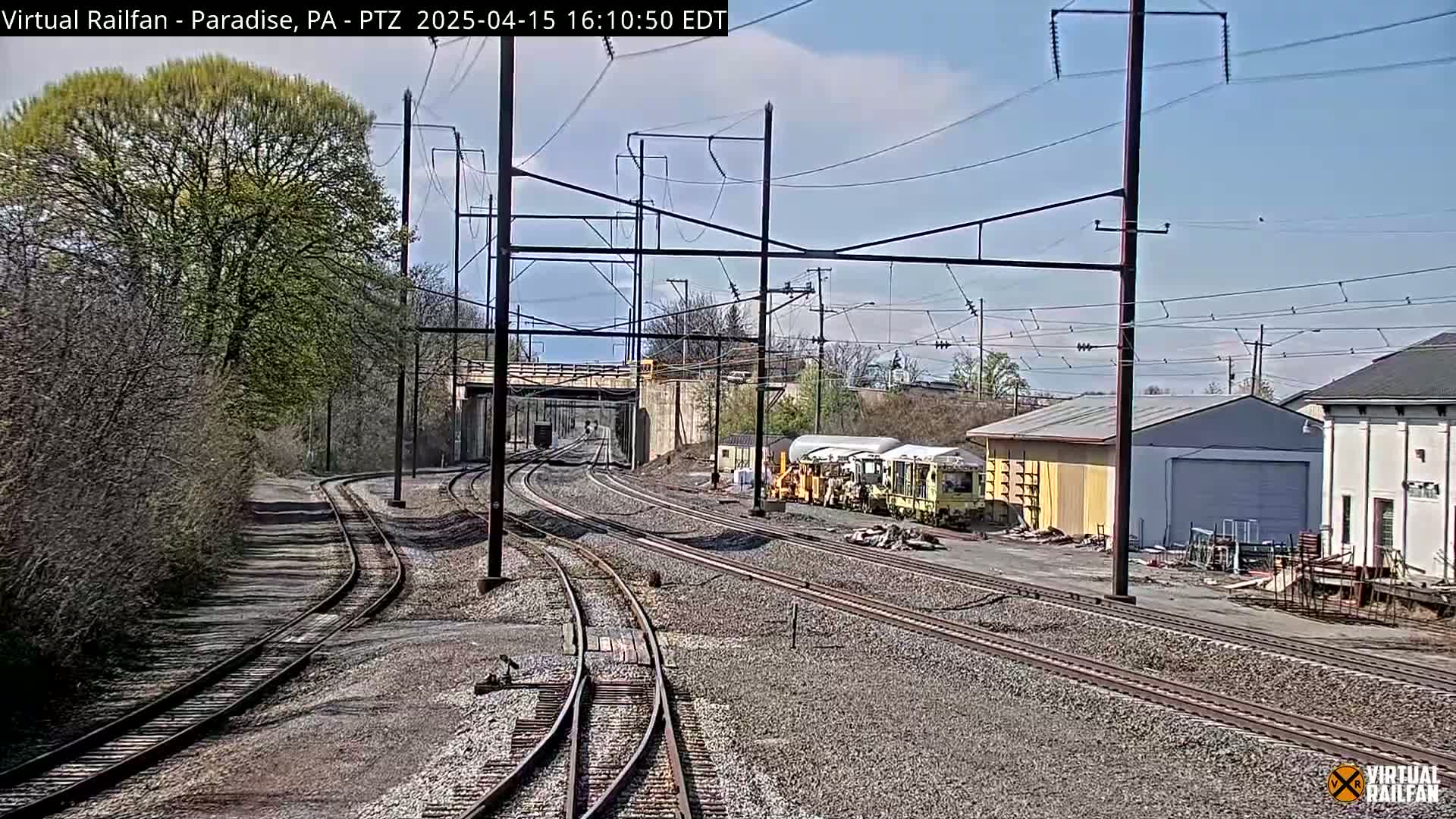 A sunny day view of several railway tracks curving around a small railway yard with some maintenance vehicles, buildings, and trees visible under a partly cloudy sky.