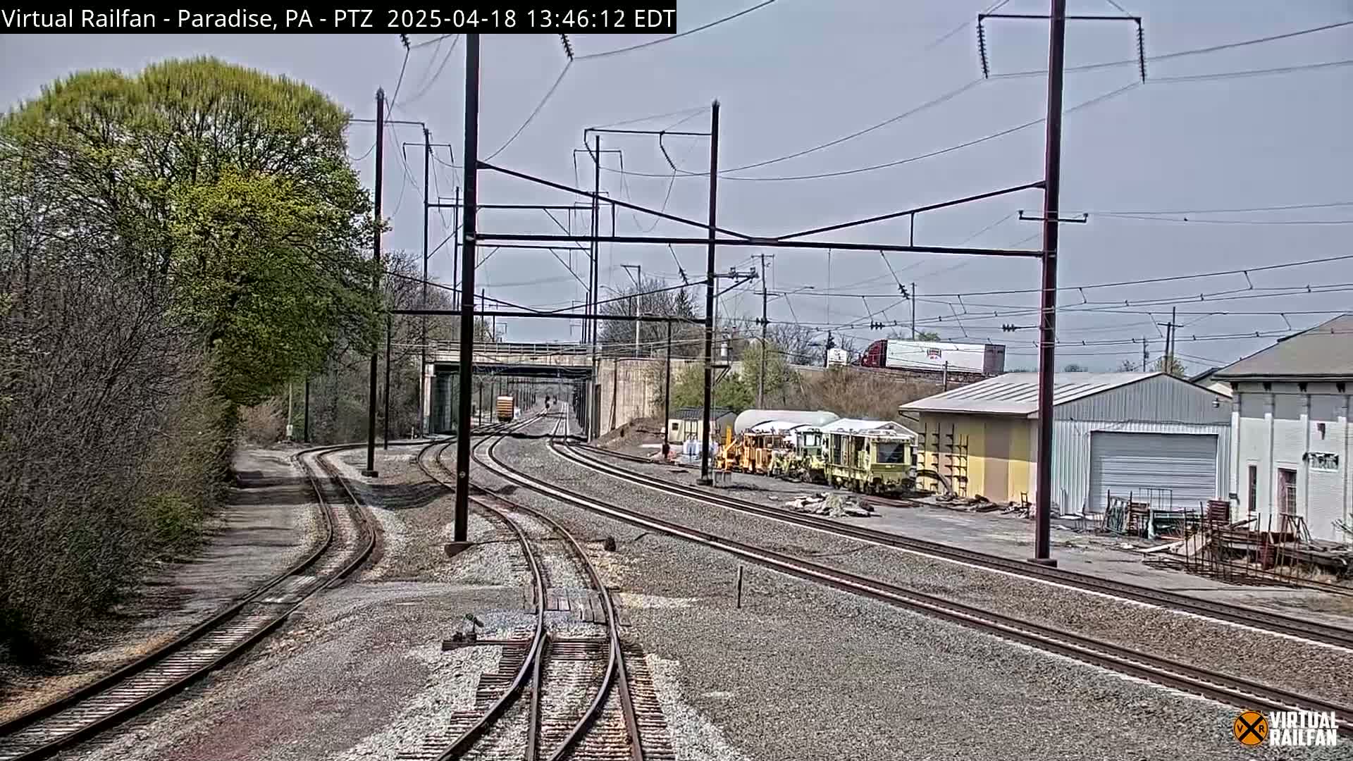 A view of railroad tracks on a partly cloudy day, with several pieces of railway maintenance equipment parked near some buildings and trees in the background.
