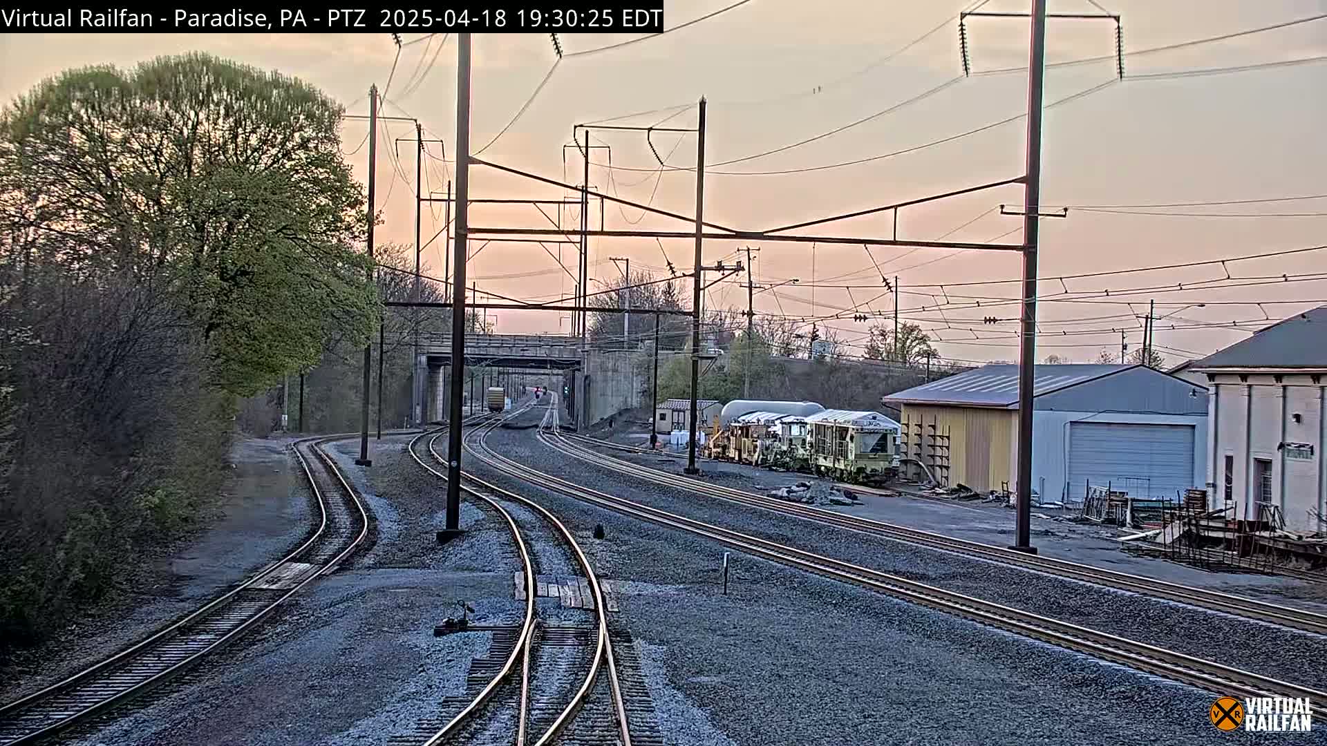 Multiple sets of train tracks converge under a pale pink twilight sky, near some buildings and greenery.