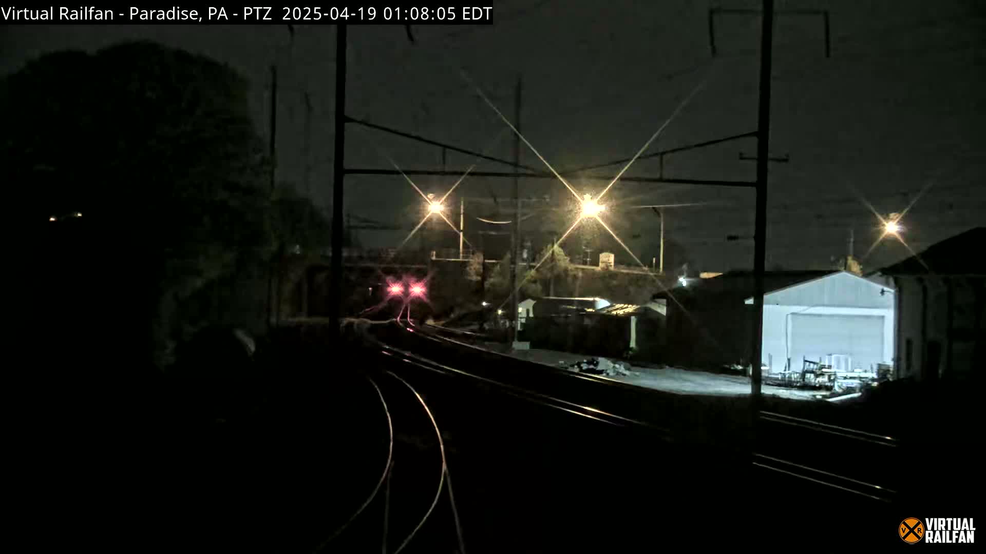 A train with illuminated red lights approaches a nighttime railway station under a dark sky.