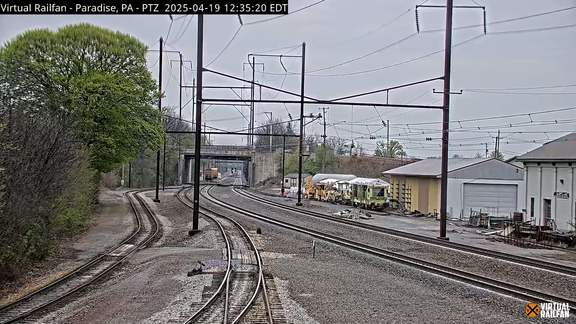 Several sets of train tracks converge near a railway yard with maintenance vehicles and buildings under an overcast sky.