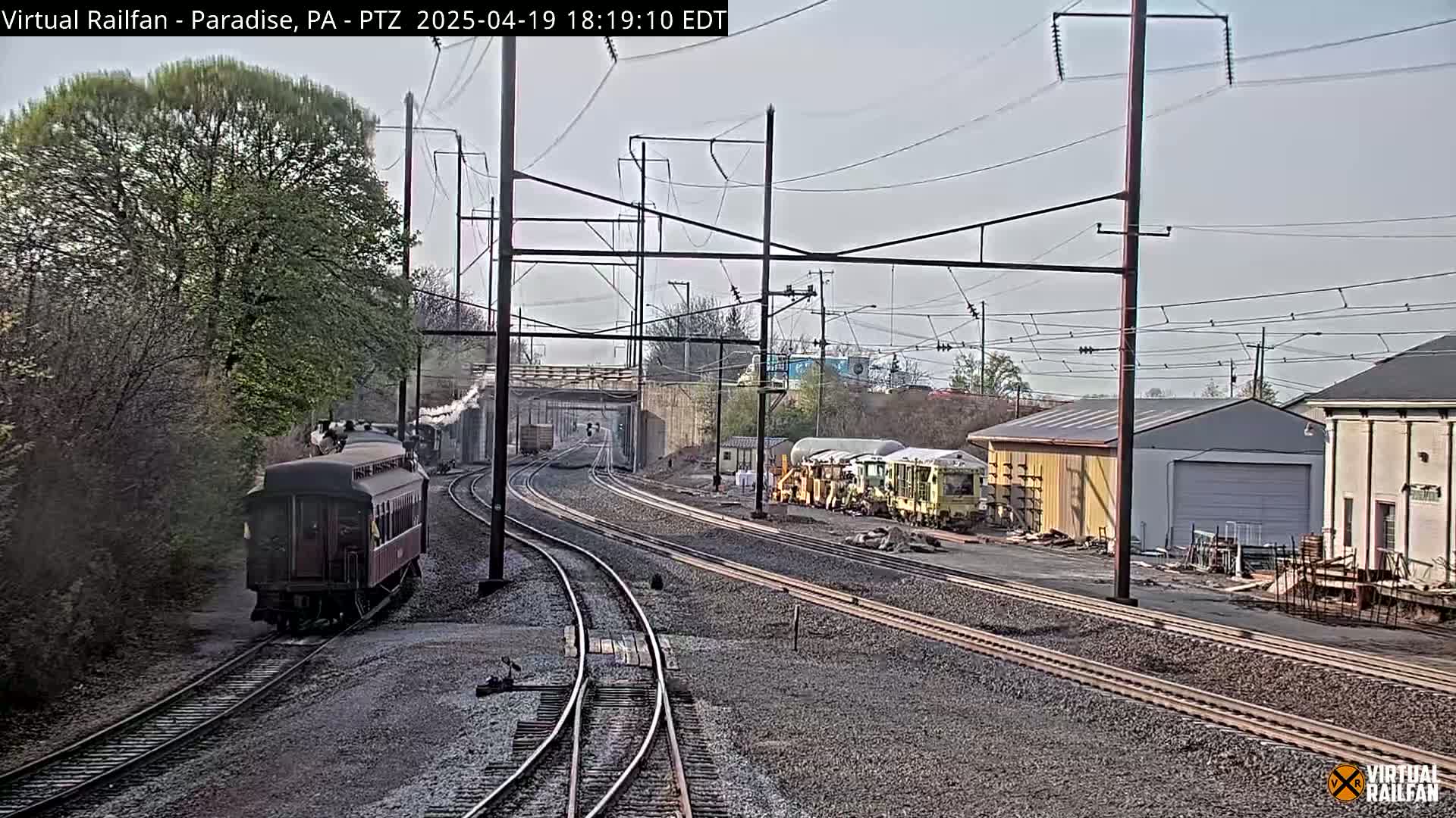 A steam train and several rail maintenance vehicles are visible on a railway track under a partly cloudy sky.