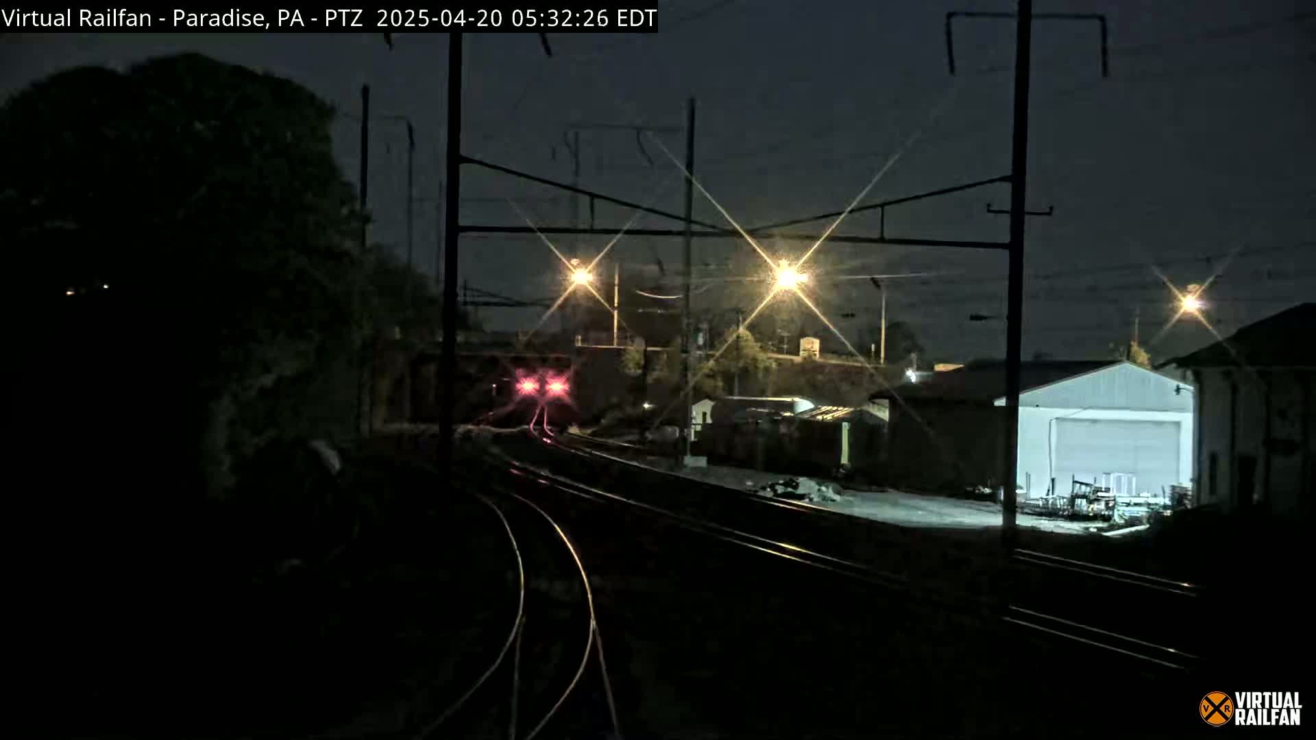 A nighttime view of a train approaching a rail yard under a dark sky illuminated by several lights.