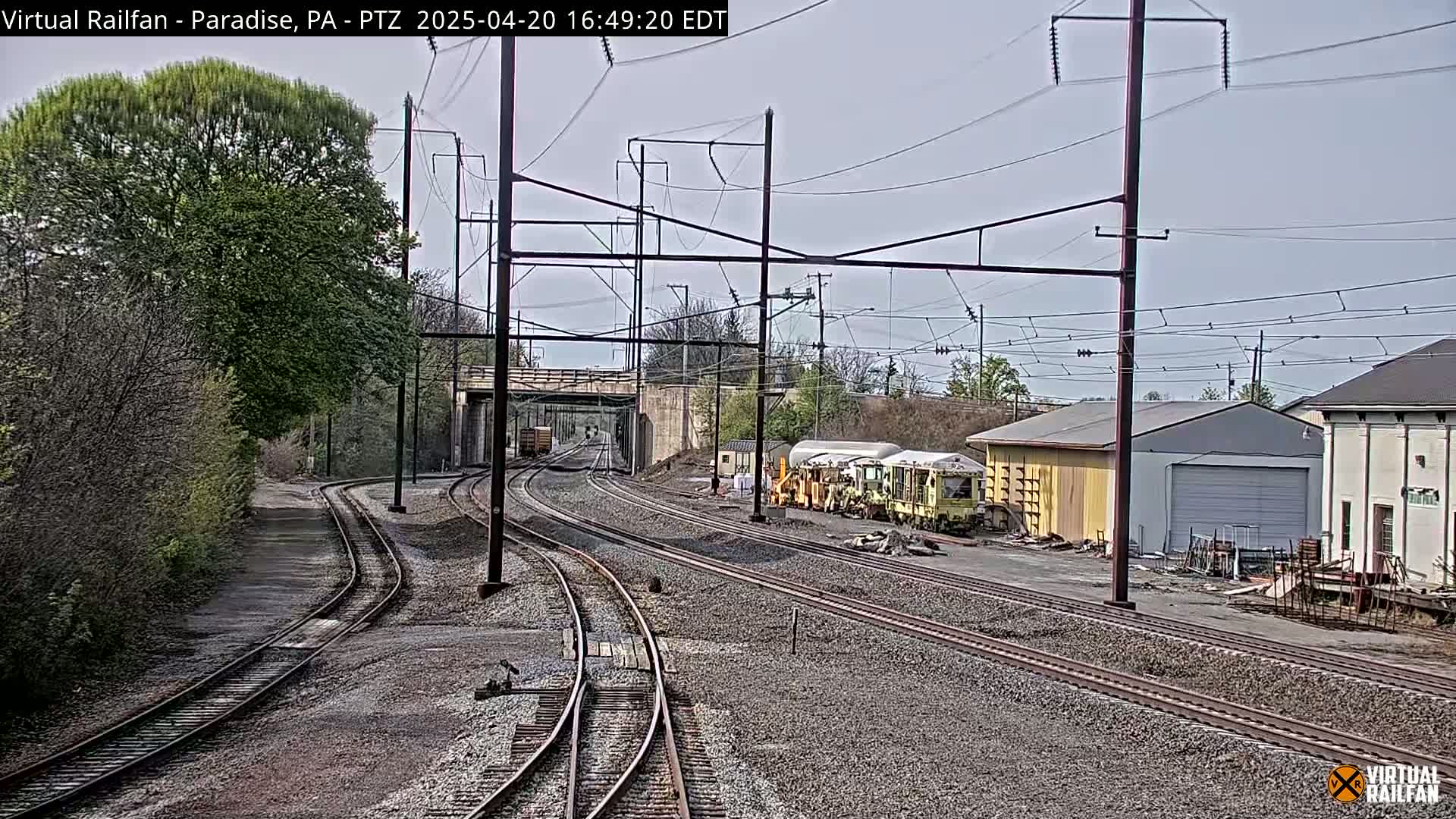 A view of railroad tracks on an overcast day, with several railcars parked near some buildings and trees bordering the sides.