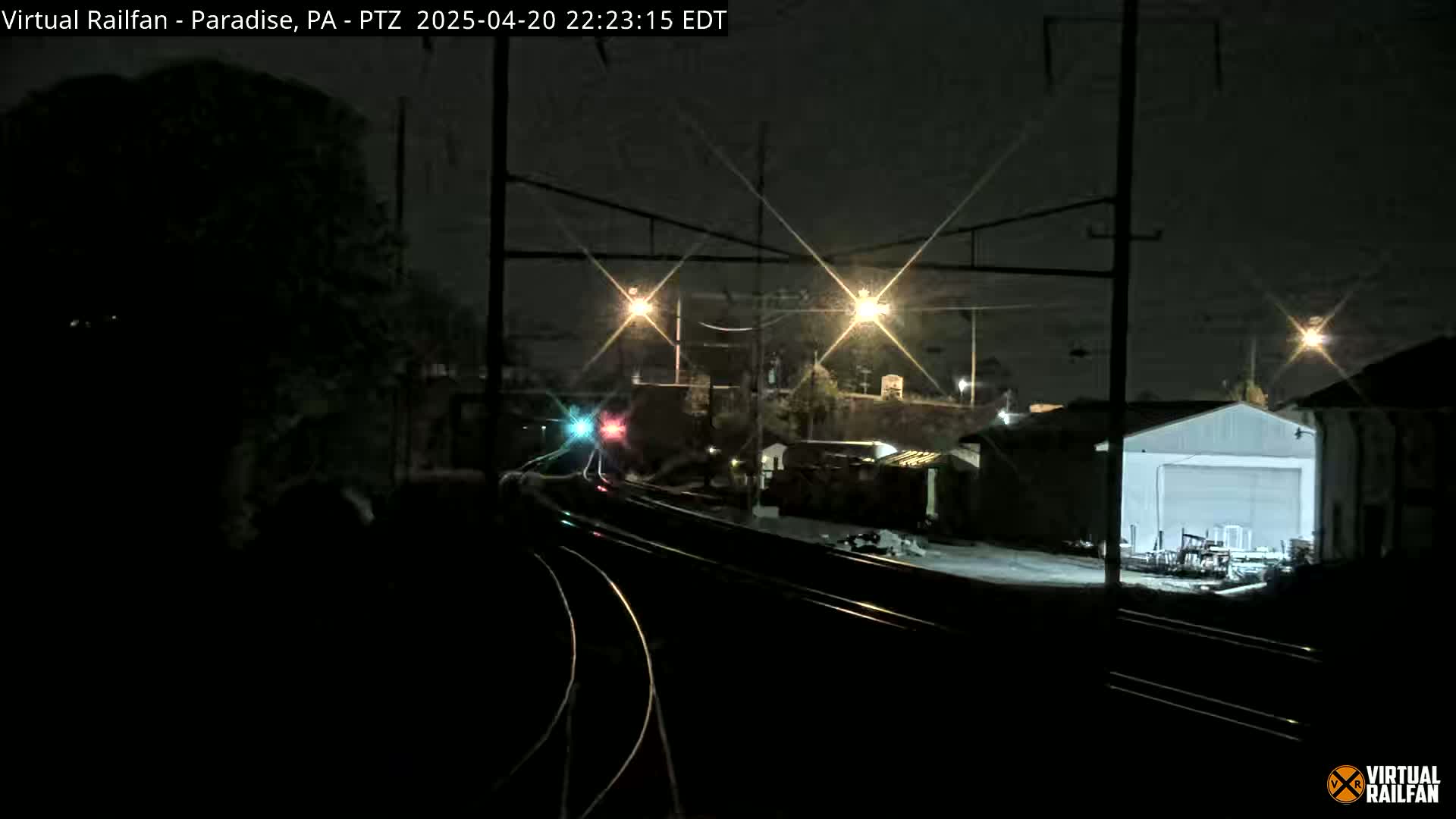 A nighttime view of a train approaching a station, with several lights illuminating the tracks and surrounding buildings.