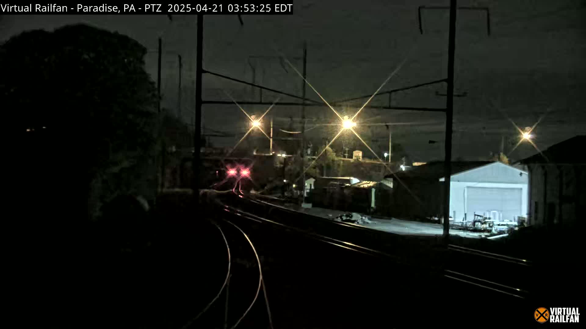 At night, a train with red lights approaches a railway junction near some buildings under a dark sky.