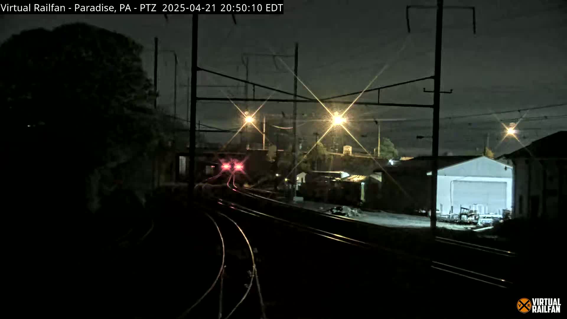 A nighttime view of a train approaching a rail yard under partly cloudy skies, illuminated by streetlights and the train's lights.