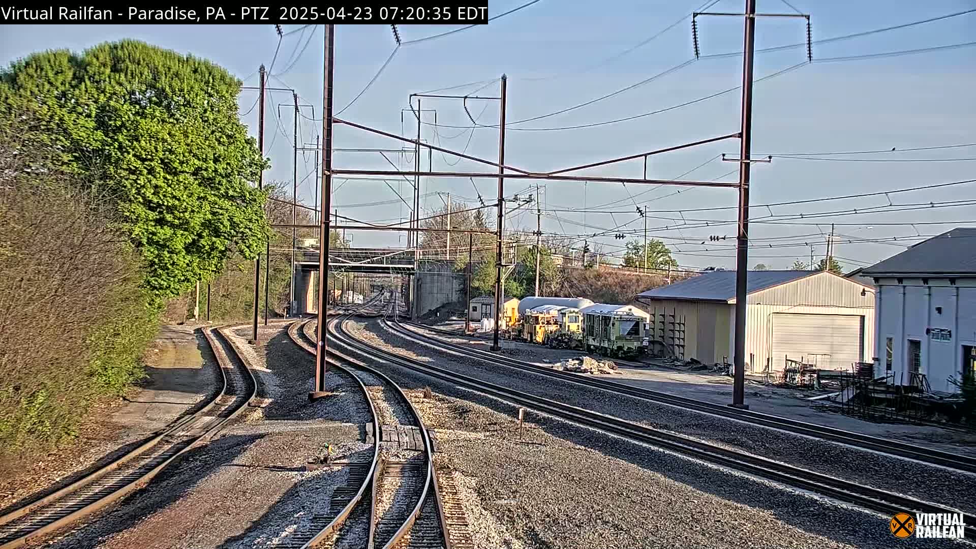 Several sets of train tracks curve toward a cluster of railway maintenance vehicles parked near some buildings under a clear, sunny sky.