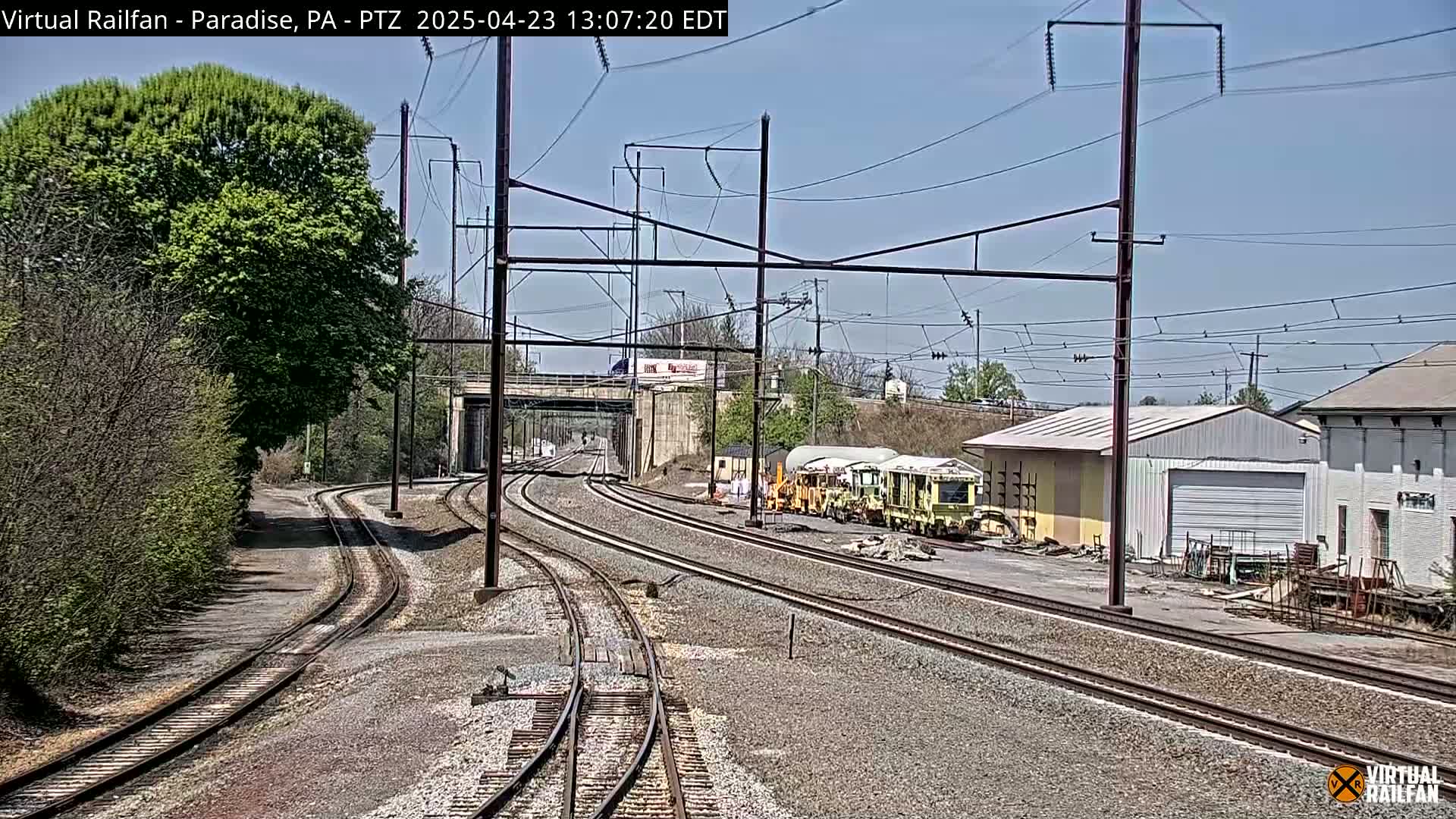 Under a clear blue sky, railroad tracks curve through a landscape of trees, shrubs, and industrial buildings, with some railway maintenance vehicles visible nearby.