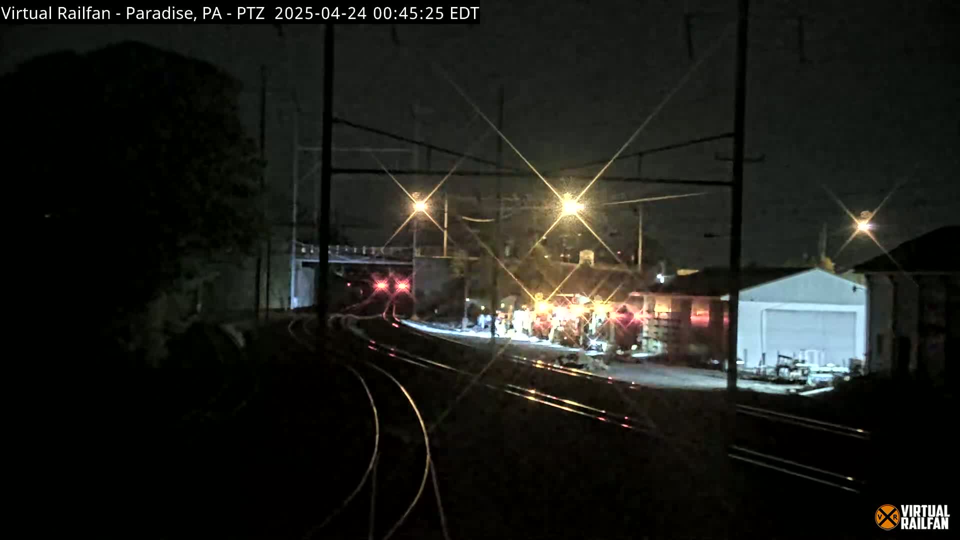 At night, under bright lights, railroad tracks wind through a dimly lit area with a building and workers near the tracks.
