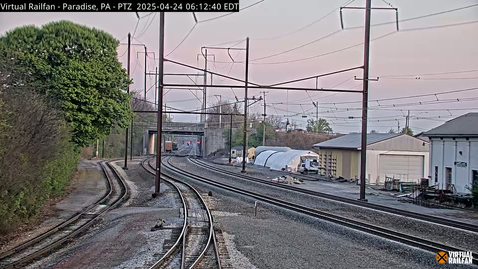 Under a twilight sky, several train tracks converge near a small industrial area with buildings and a temporary structure, alongside trees and vegetation.