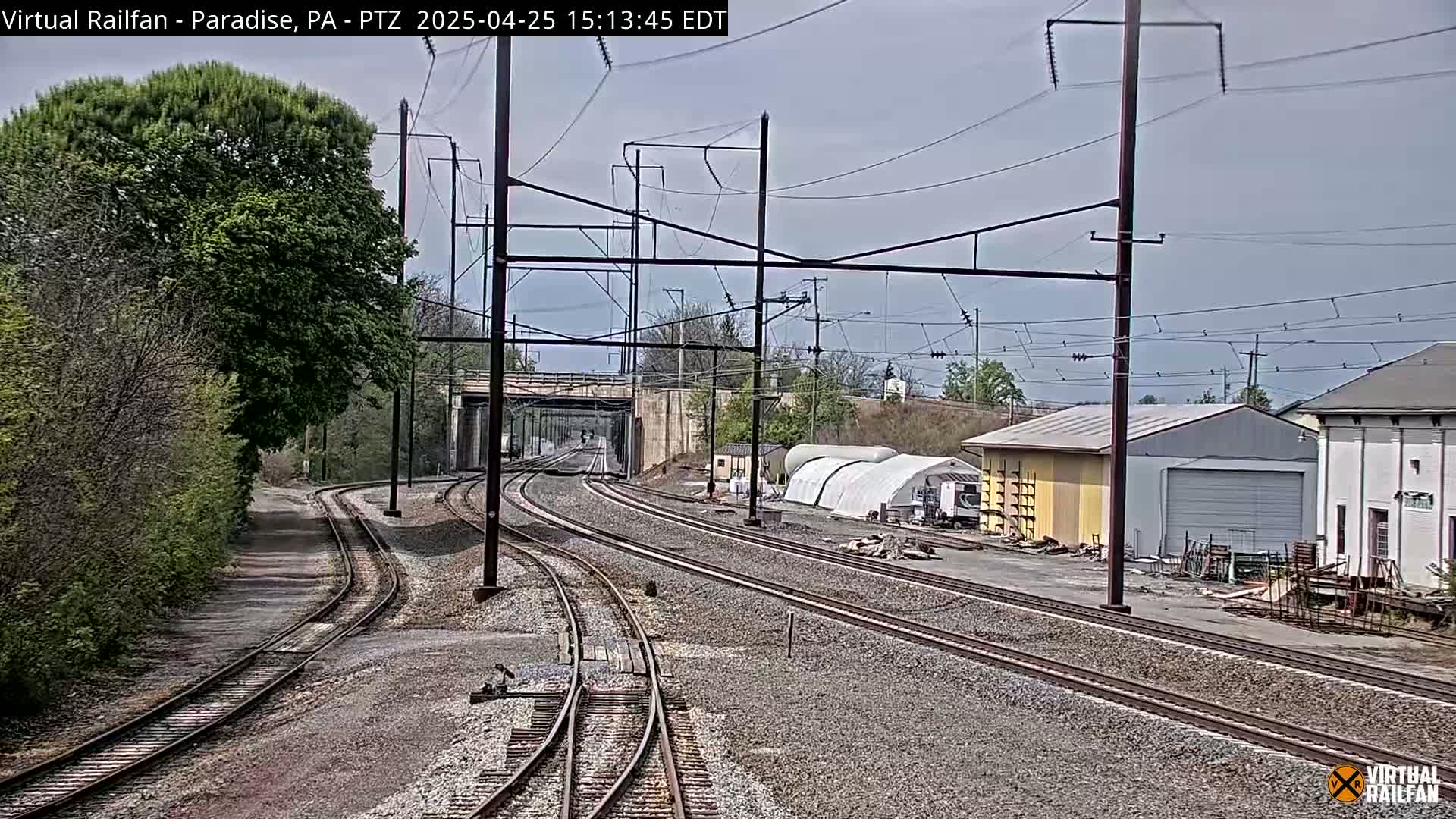 Multiple sets of train tracks converge near a railway underpass under a partly cloudy sky, with buildings and trees lining the sides.