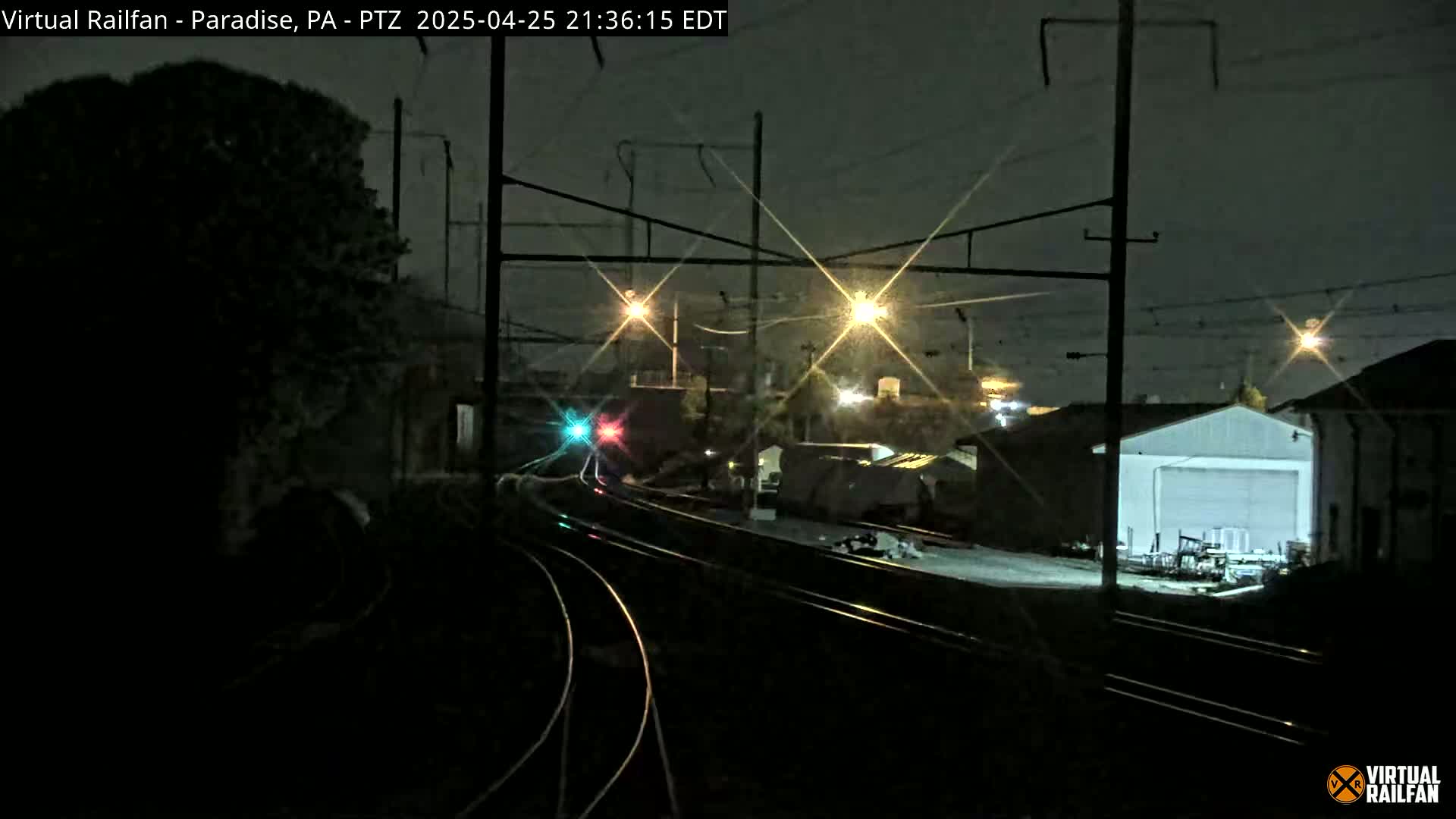 At night, a nighttime view shows railroad tracks leading to a building under bright lights, with a train's lights visible in the distance.
