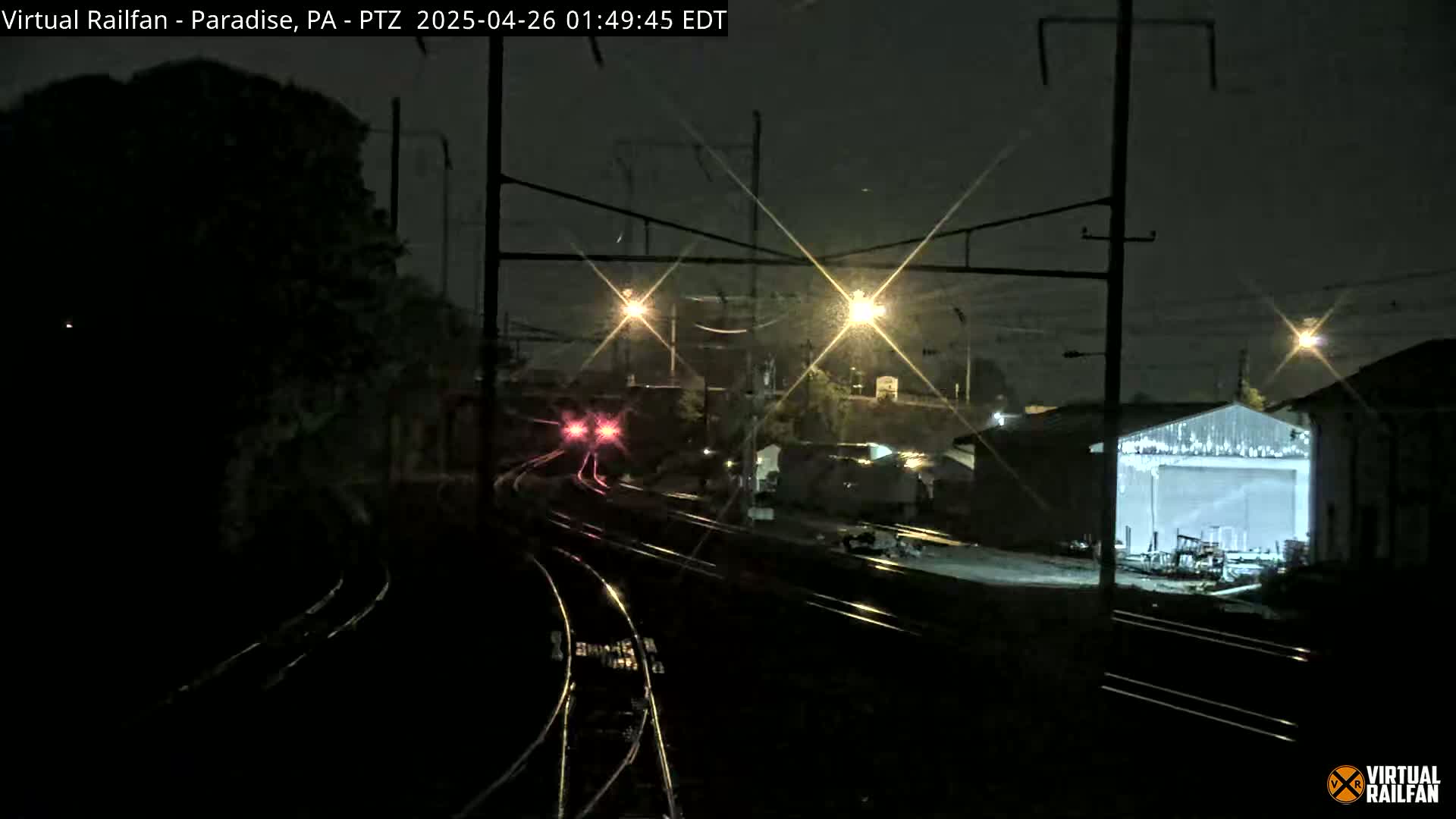A nighttime view of a train passing by several buildings and railroad tracks under a dark, possibly rainy sky.