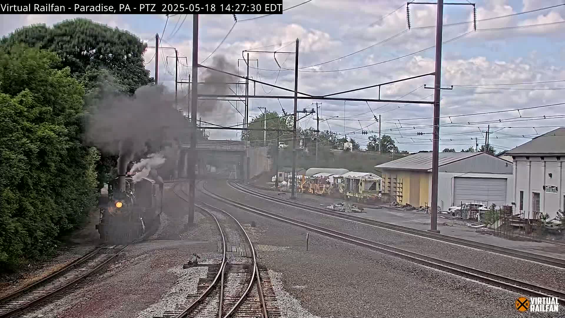 A steam train, emitting a large plume of smoke, approaches a railway junction under a partly cloudy sky.