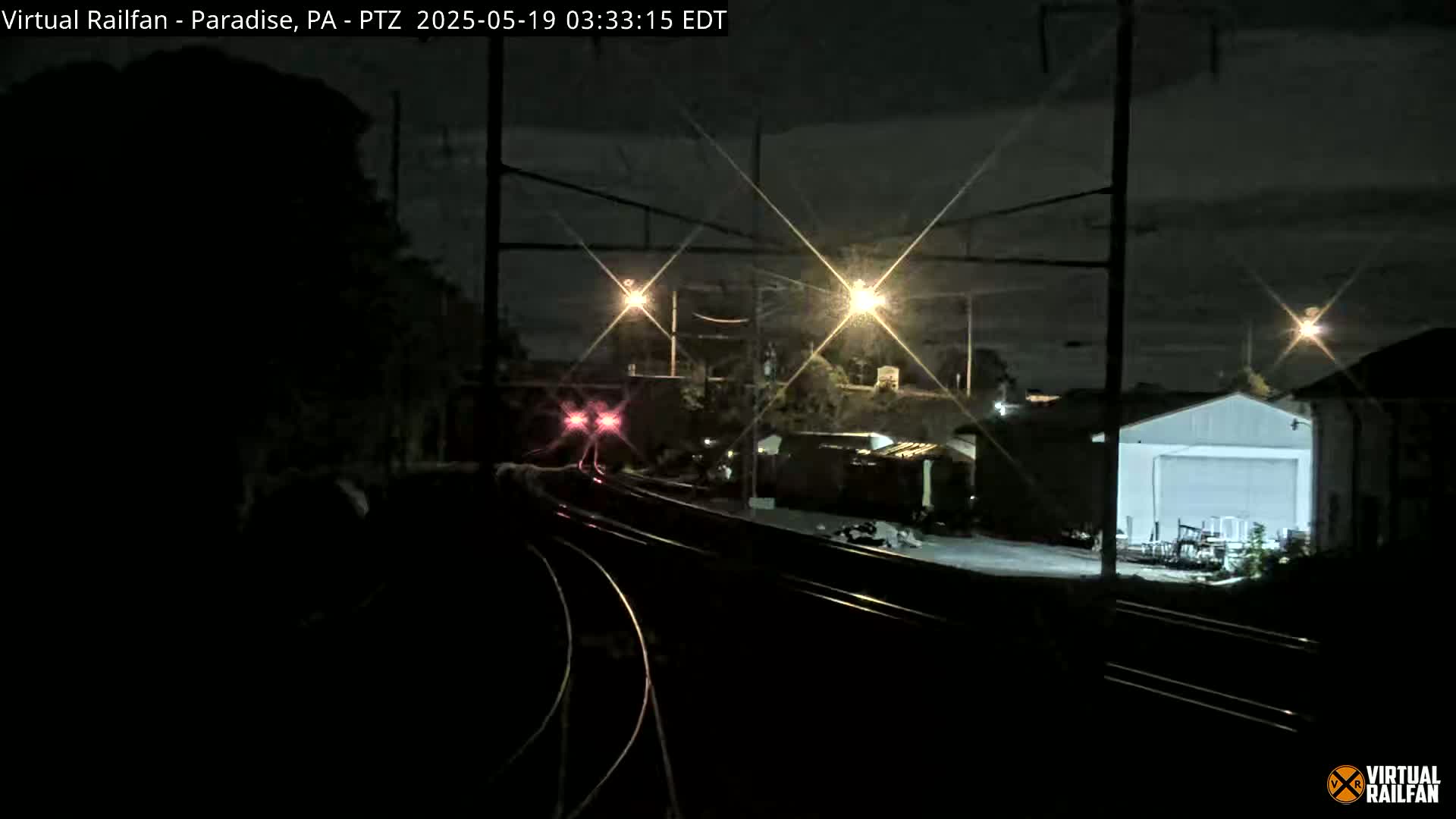A nighttime view of railroad tracks with a train approaching, passing by buildings under a partly cloudy sky illuminated by streetlights.