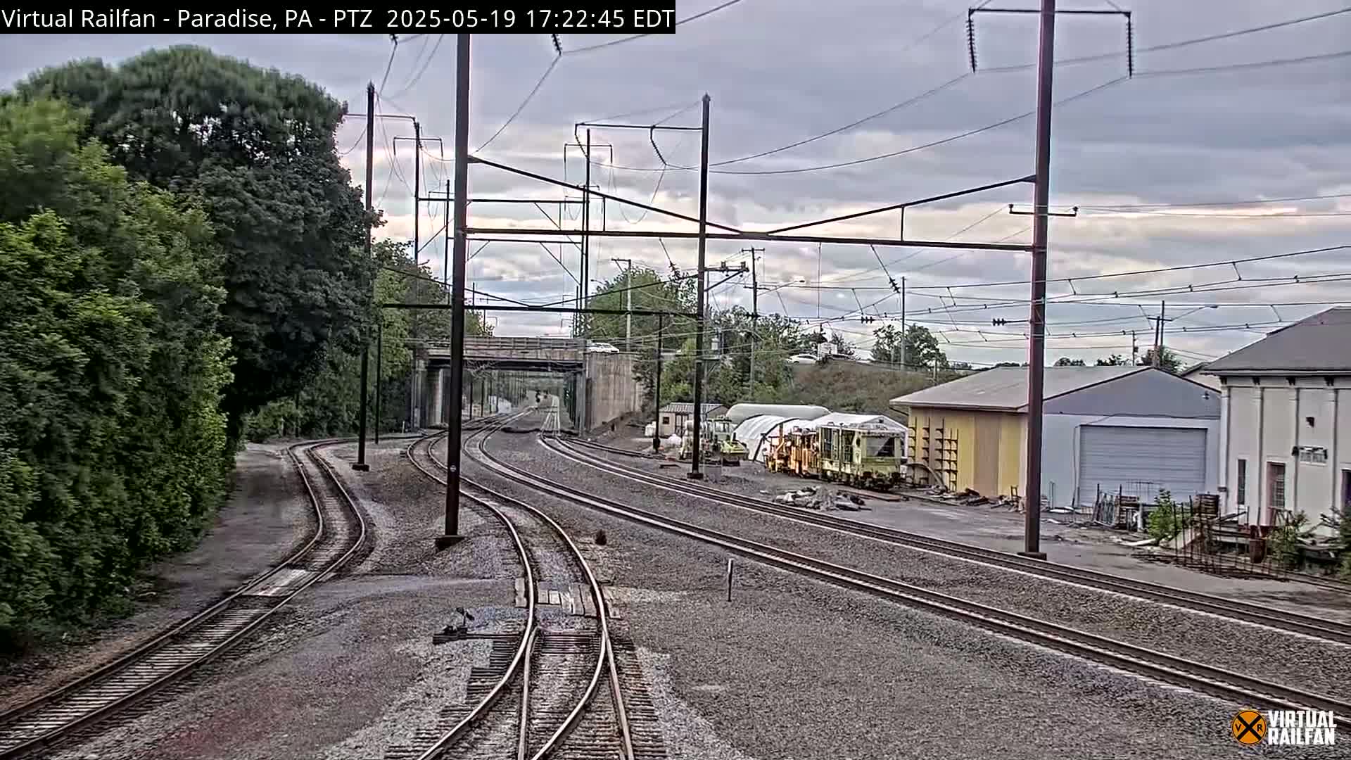 Multiple train tracks diverge near a roadway underpass, past industrial buildings and lush green trees under an overcast sky.