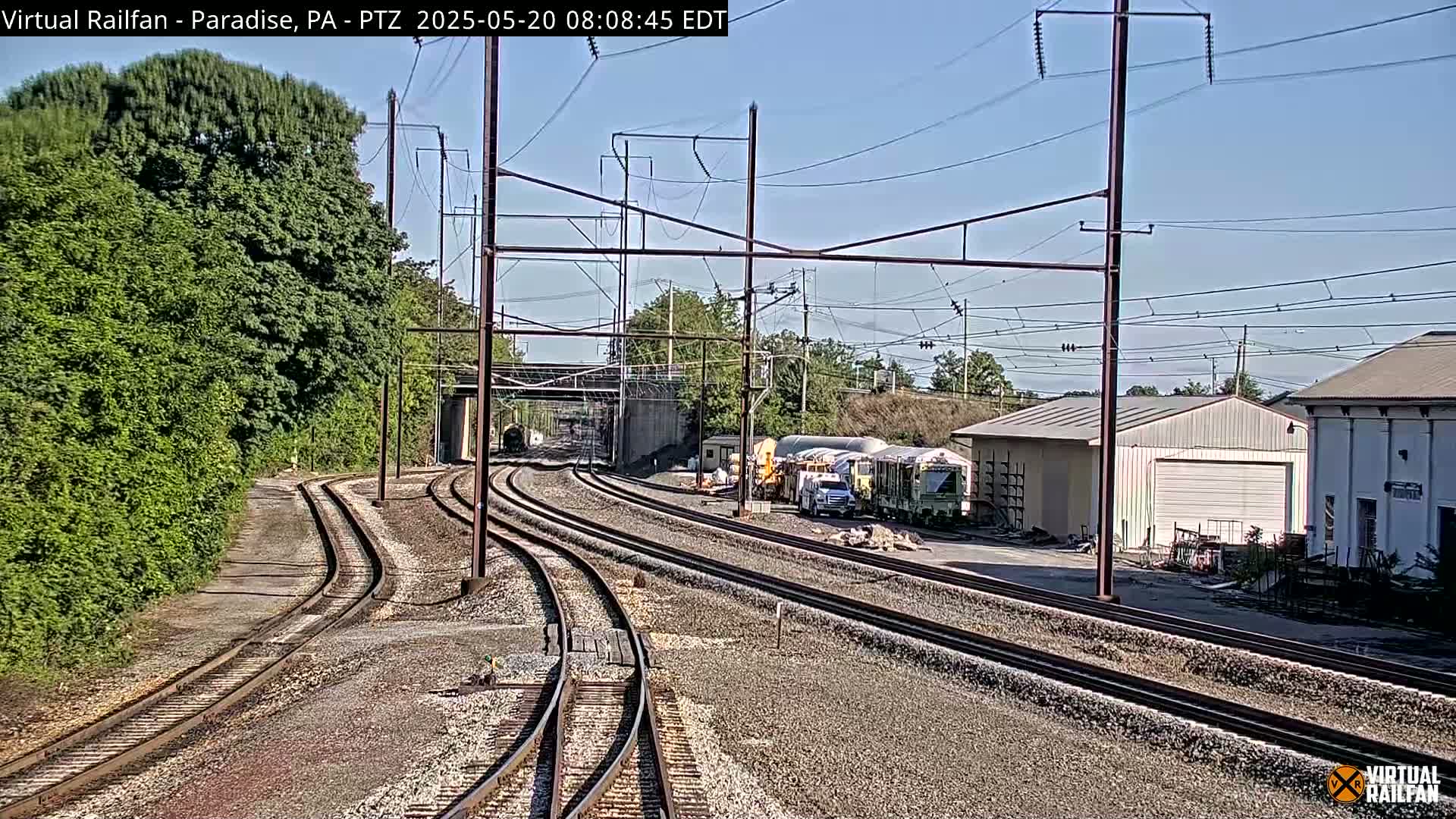 Several sets of train tracks curve toward a rail yard containing industrial buildings and railway maintenance equipment under a clear sky.