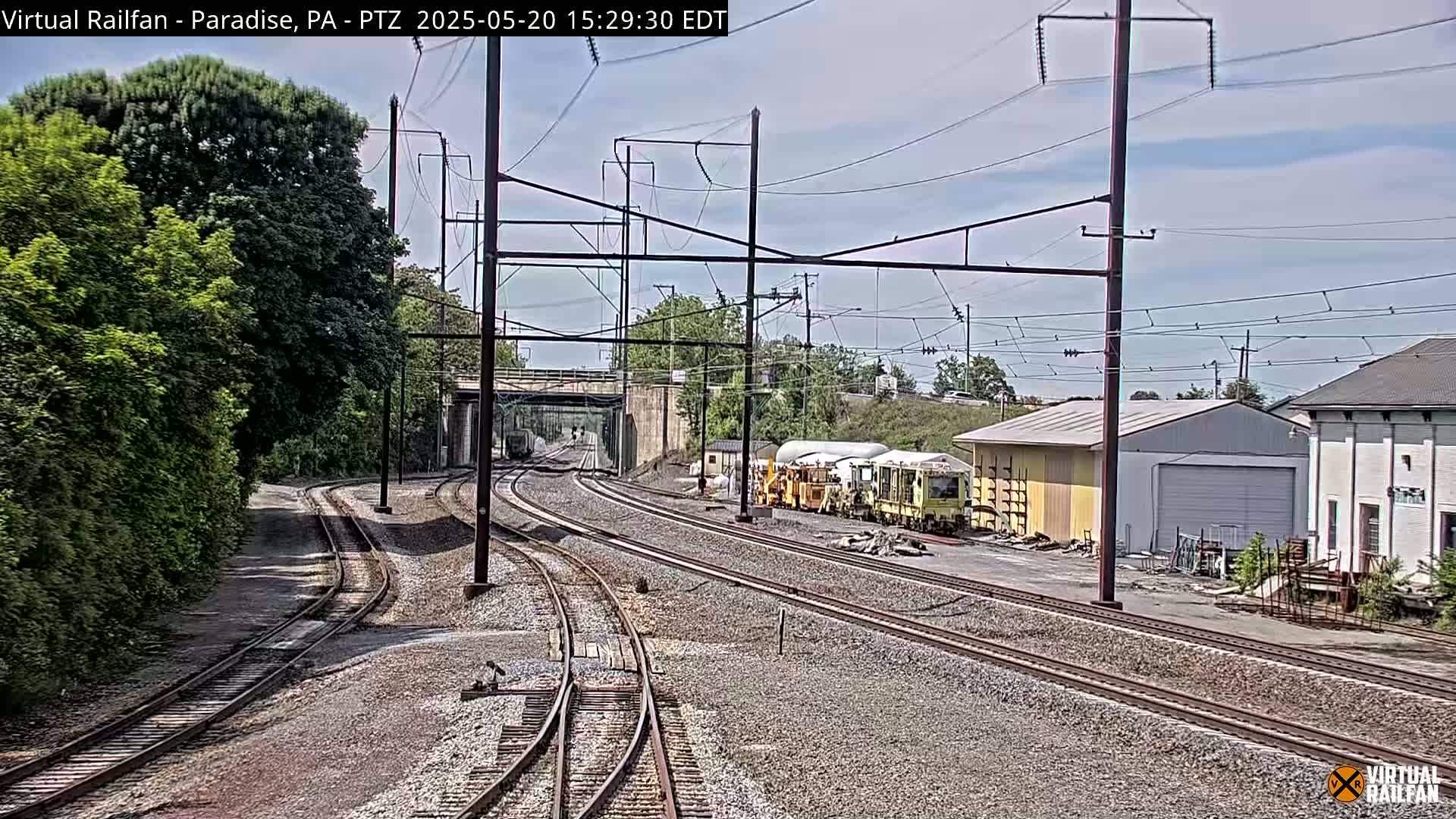 Under a partly cloudy sky, several railway tracks converge near industrial buildings and lush green vegetation.
