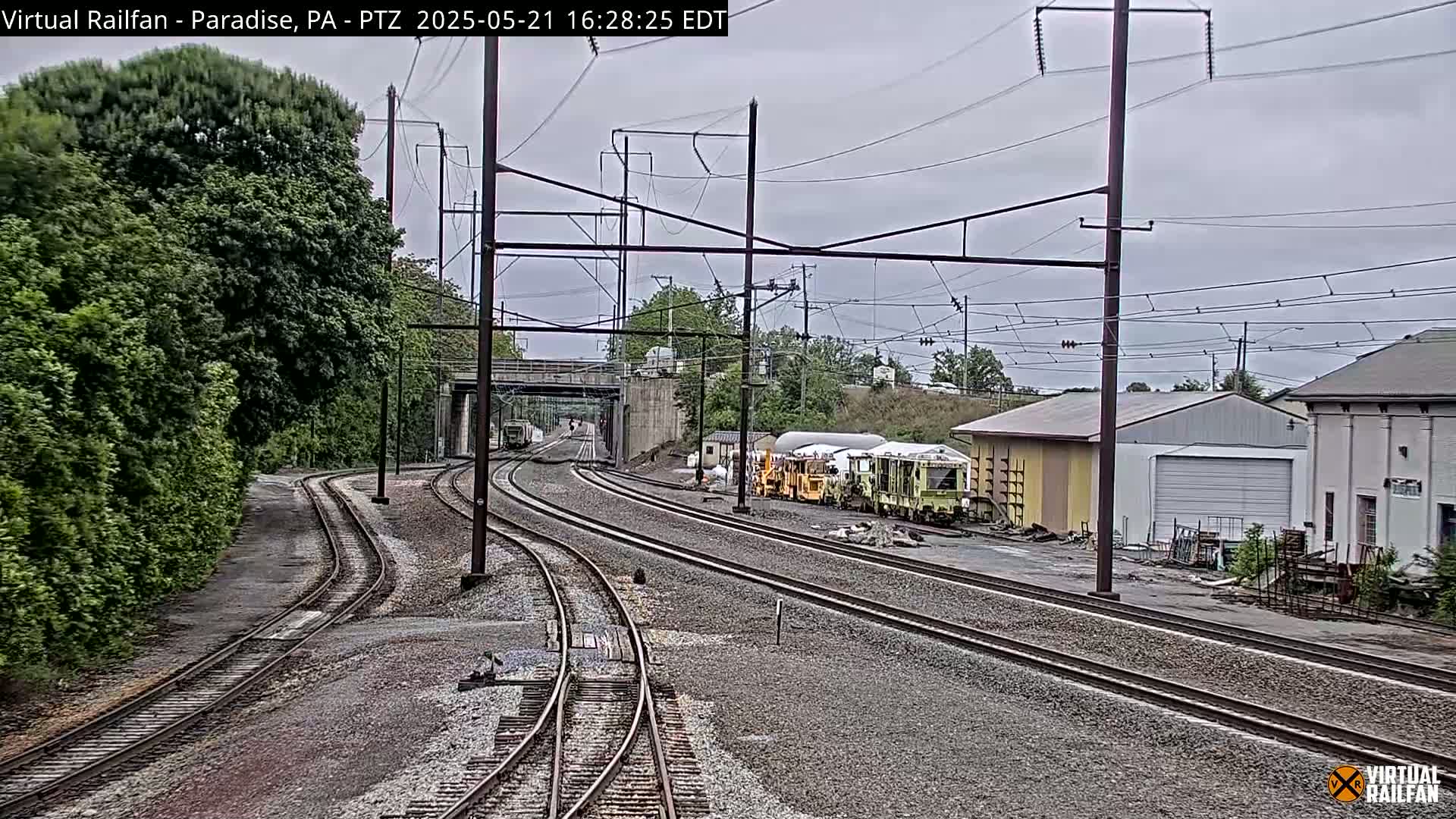 Several sets of train tracks converge near some railway maintenance vehicles parked beside industrial buildings under an overcast sky.