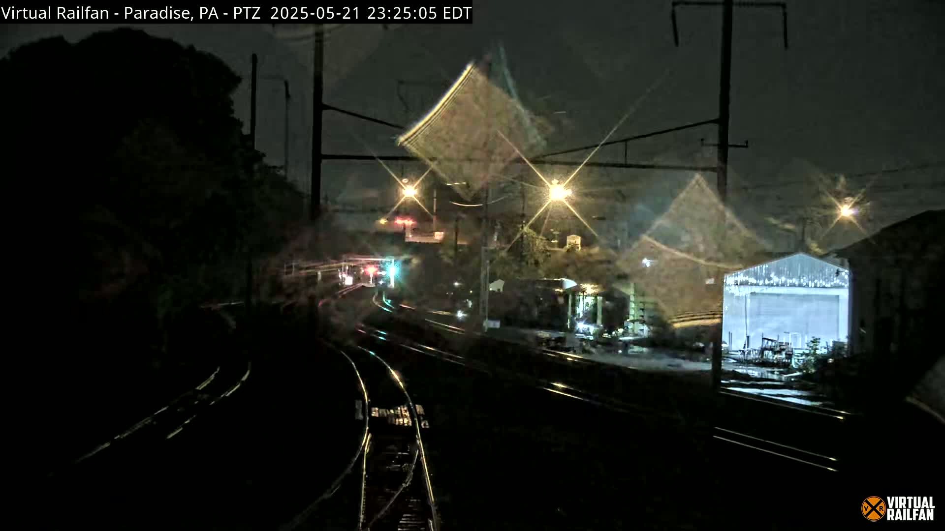 A nighttime view of rain-slicked train tracks curving through a dimly lit area with buildings and trees, under streetlights.