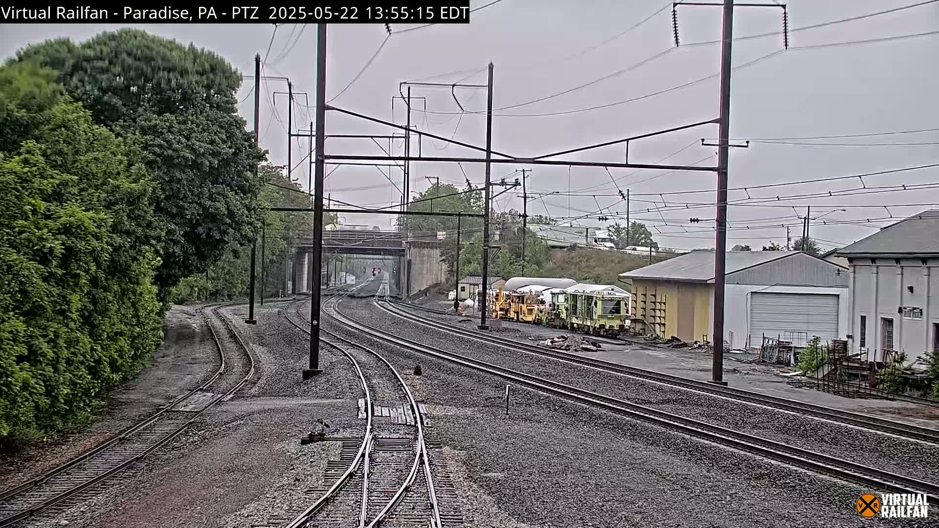 Several sets of train tracks diverge from a central point, passing under a bridge and toward some buildings and railway maintenance vehicles under an overcast sky.