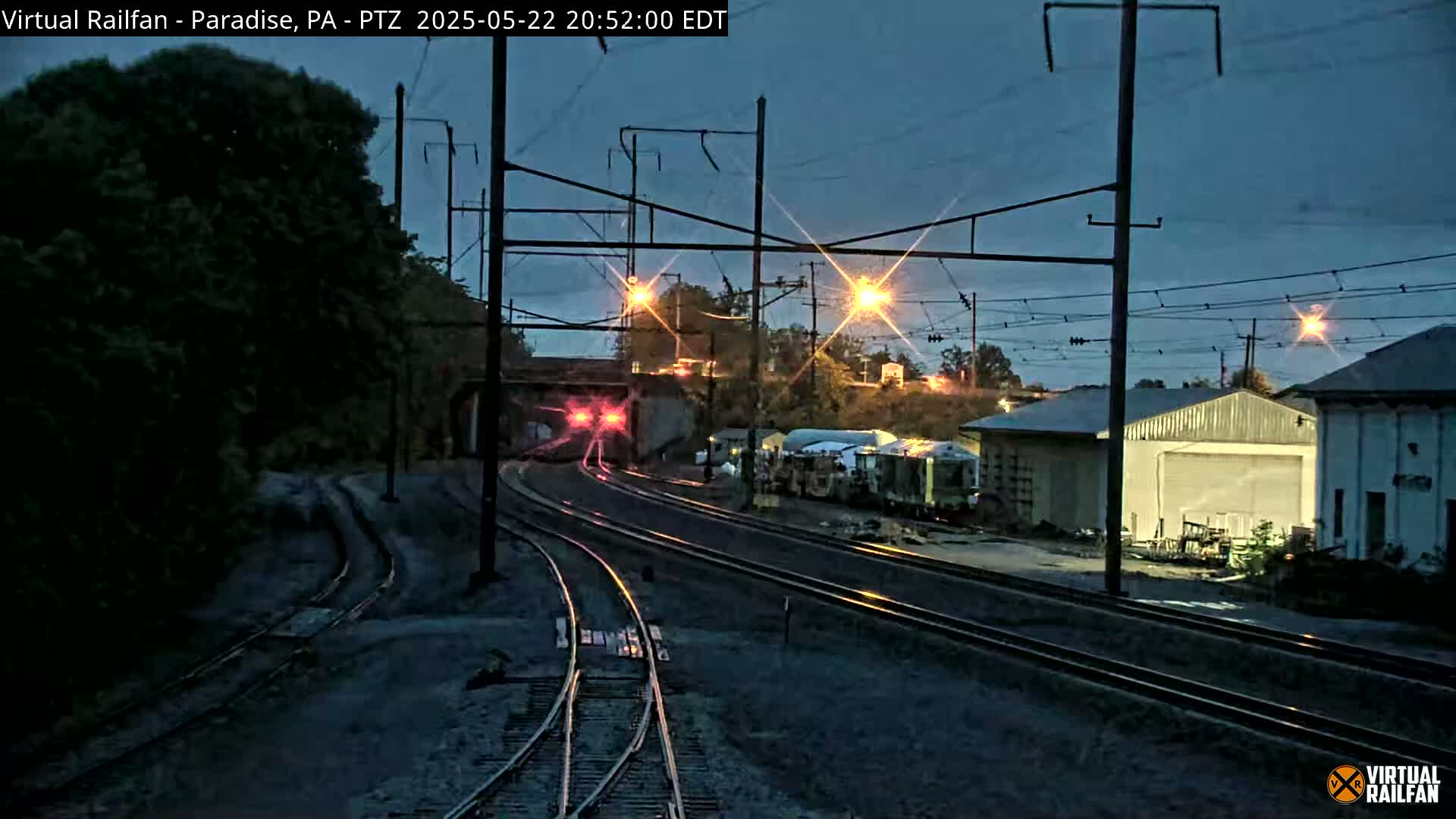 A nighttime view of a train yard with several sets of railroad tracks, some buildings, and a train partially visible exiting a tunnel under a bridge.