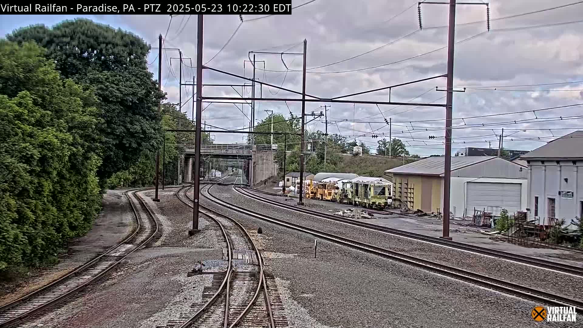 Multiple train tracks curve through a landscape of green trees, a small industrial area with buildings, and an overcast sky.