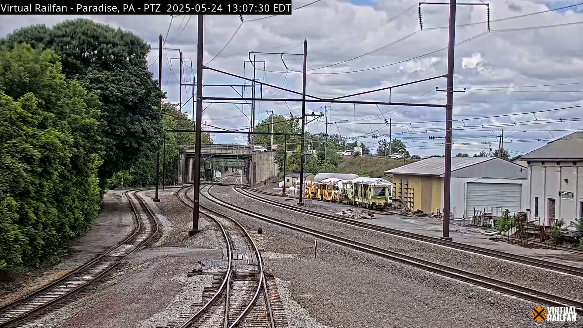Multiple sets of train tracks converge near some buildings and green trees under a partly cloudy sky.