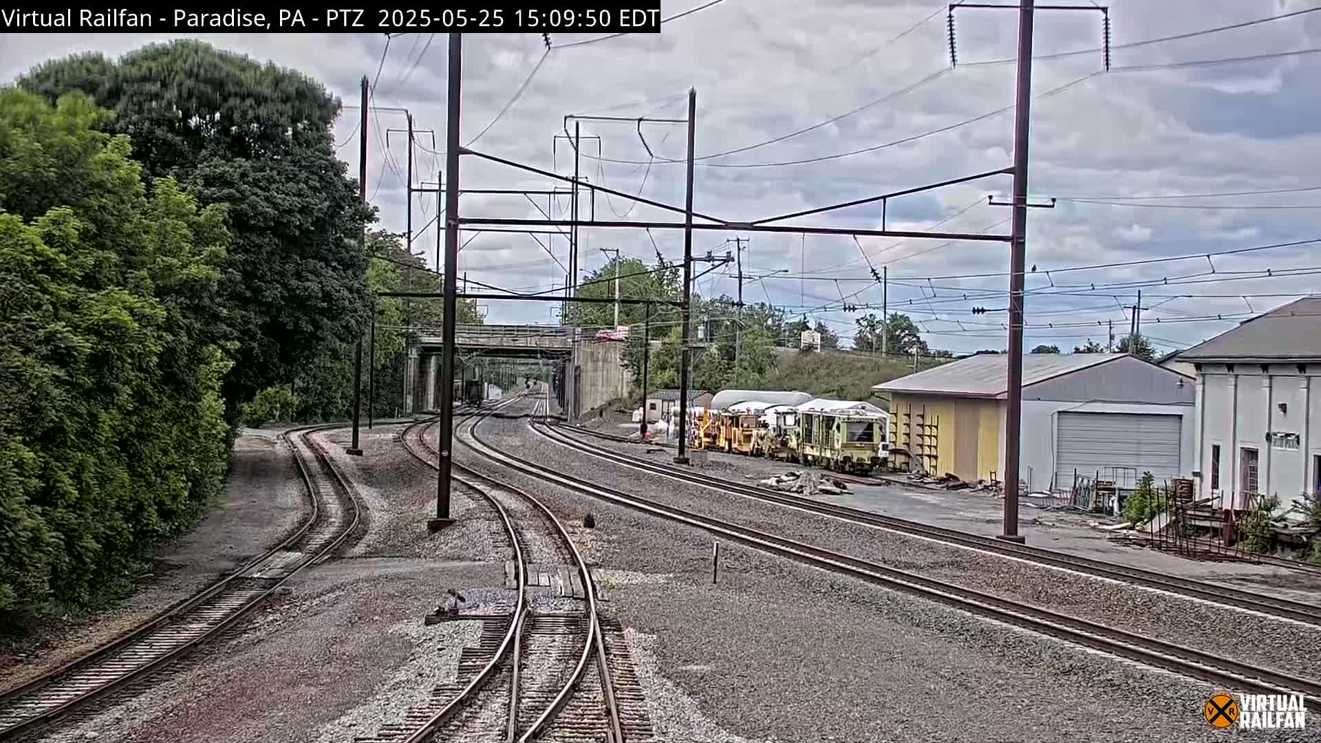 Several sets of train tracks converge near a rail yard containing industrial buildings under a cloudy sky.