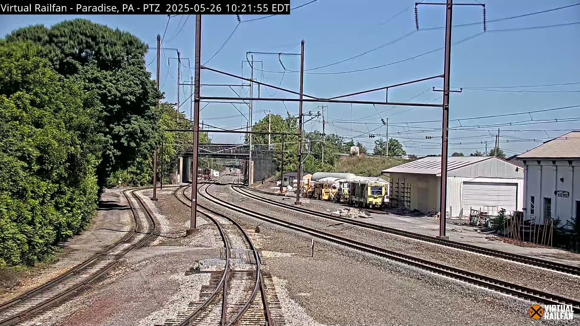 A sunny day at a railway yard with multiple train tracks converging, several railway cars parked near buildings, and lush green vegetation bordering the scene.