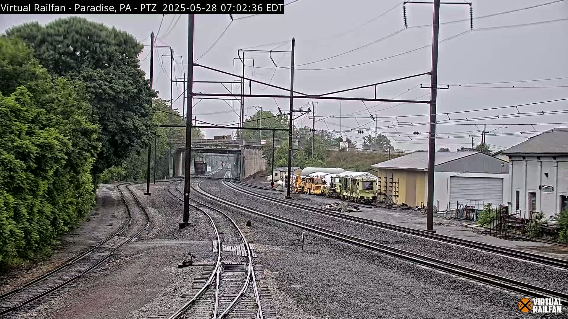 Several sets of train tracks curve through a landscape of green trees and industrial buildings under an overcast sky.