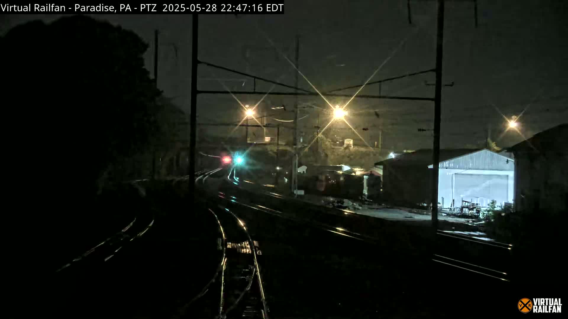 A nighttime view of a train yard with rain, showing railroad tracks, a train in the distance, and some buildings under lights.