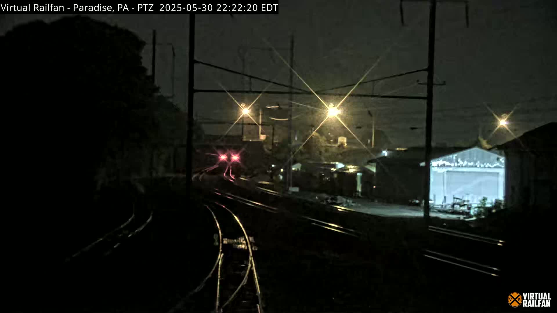 A nighttime view of railroad tracks and a train approaching a station, illuminated by streetlights under a dark sky.