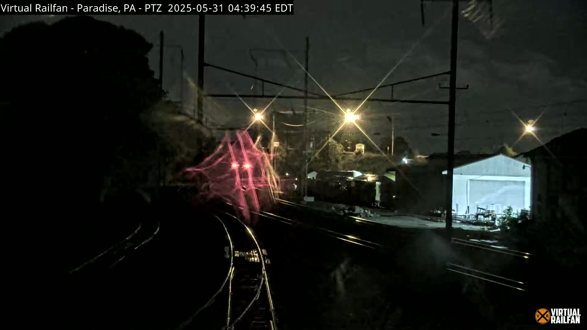 A nighttime view shows a train passing by railroad tracks near buildings under a partly cloudy sky.