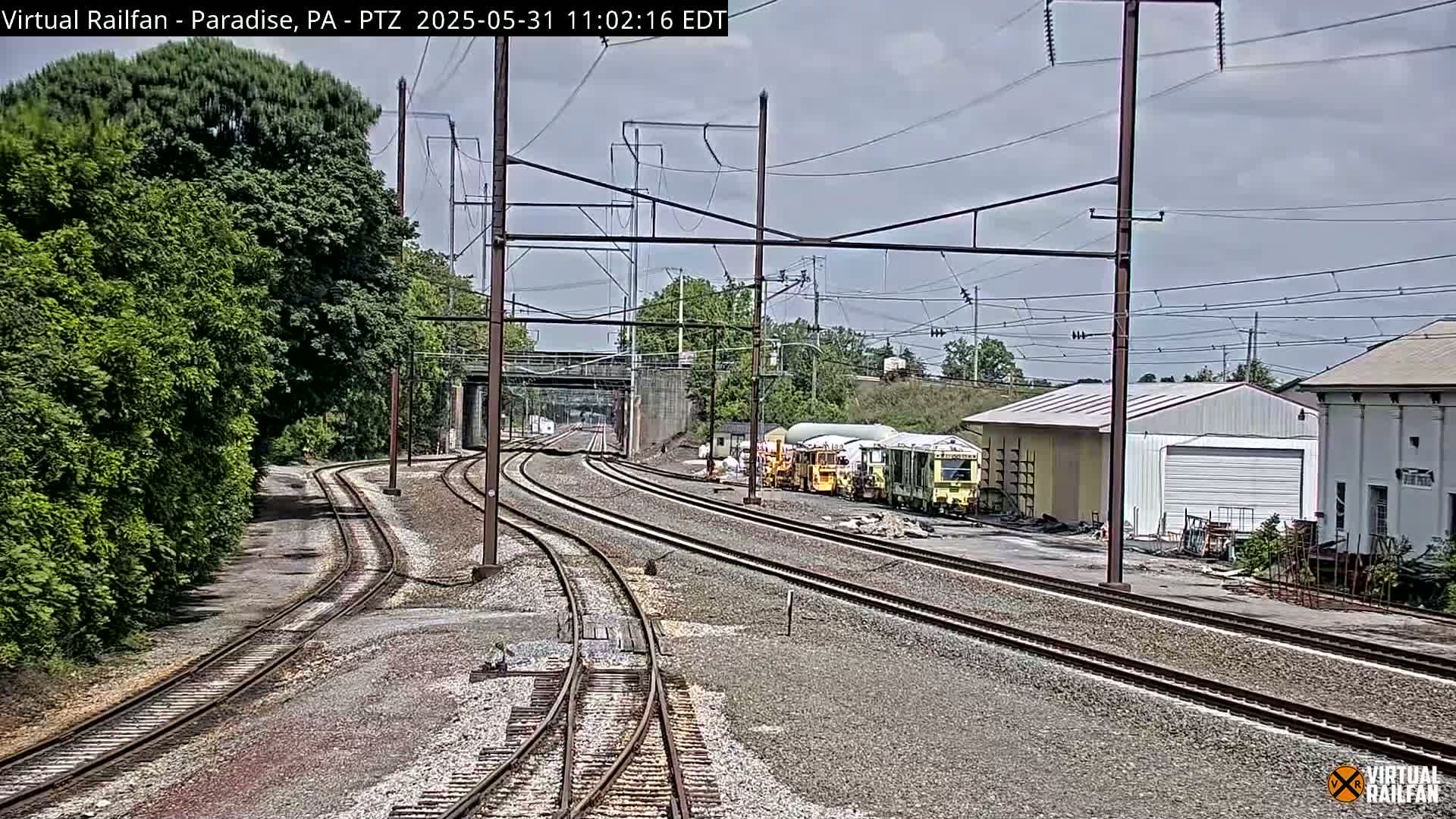 Several sets of train tracks curve through a yard with several railcars parked near a few buildings under an overcast sky.