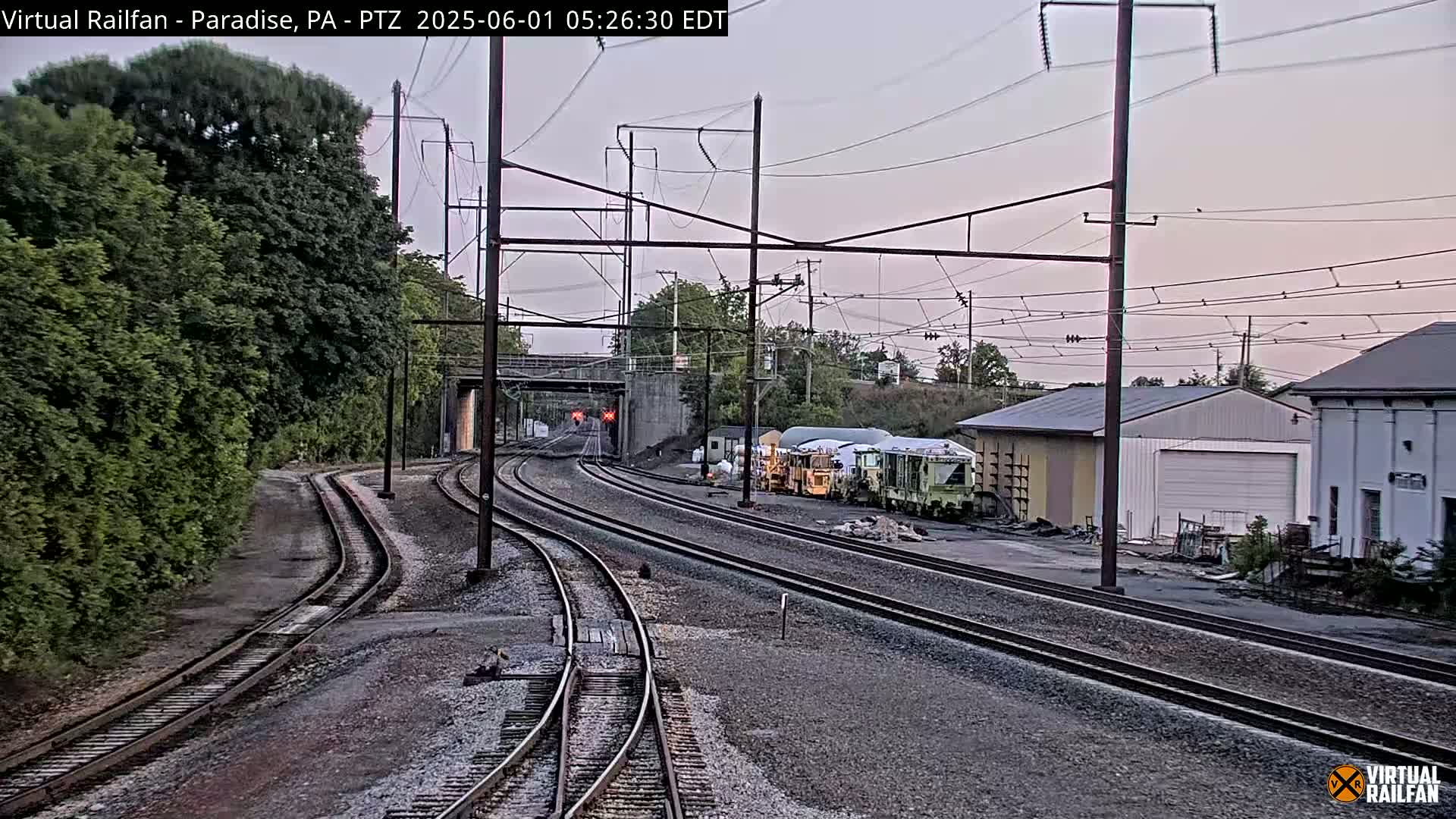 Several sets of train tracks converge near a railway yard containing several pieces of railway equipment under a twilight sky.