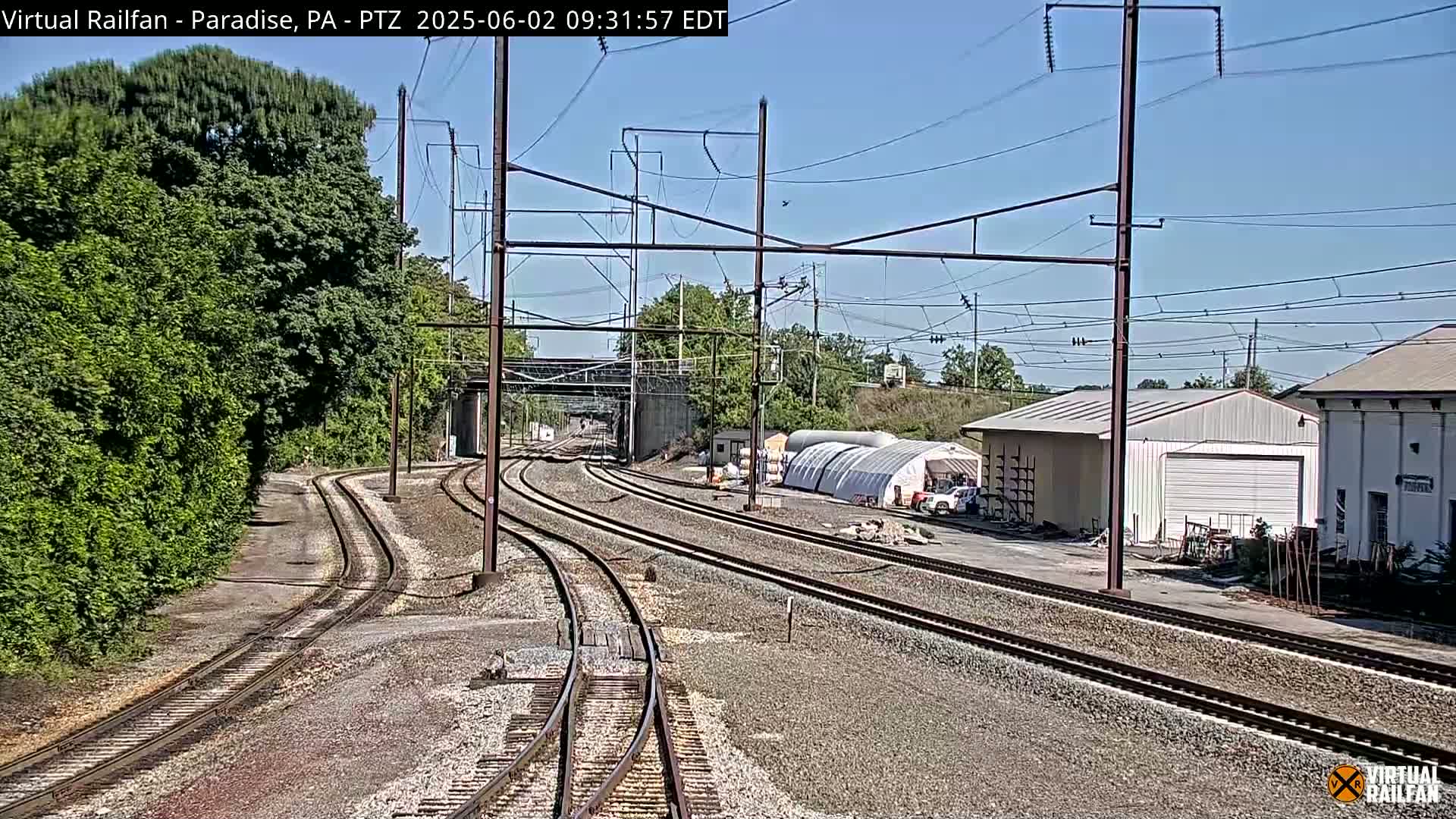 Several sets of train tracks curve gently away from the viewer, past a small industrial area on a sunny day.