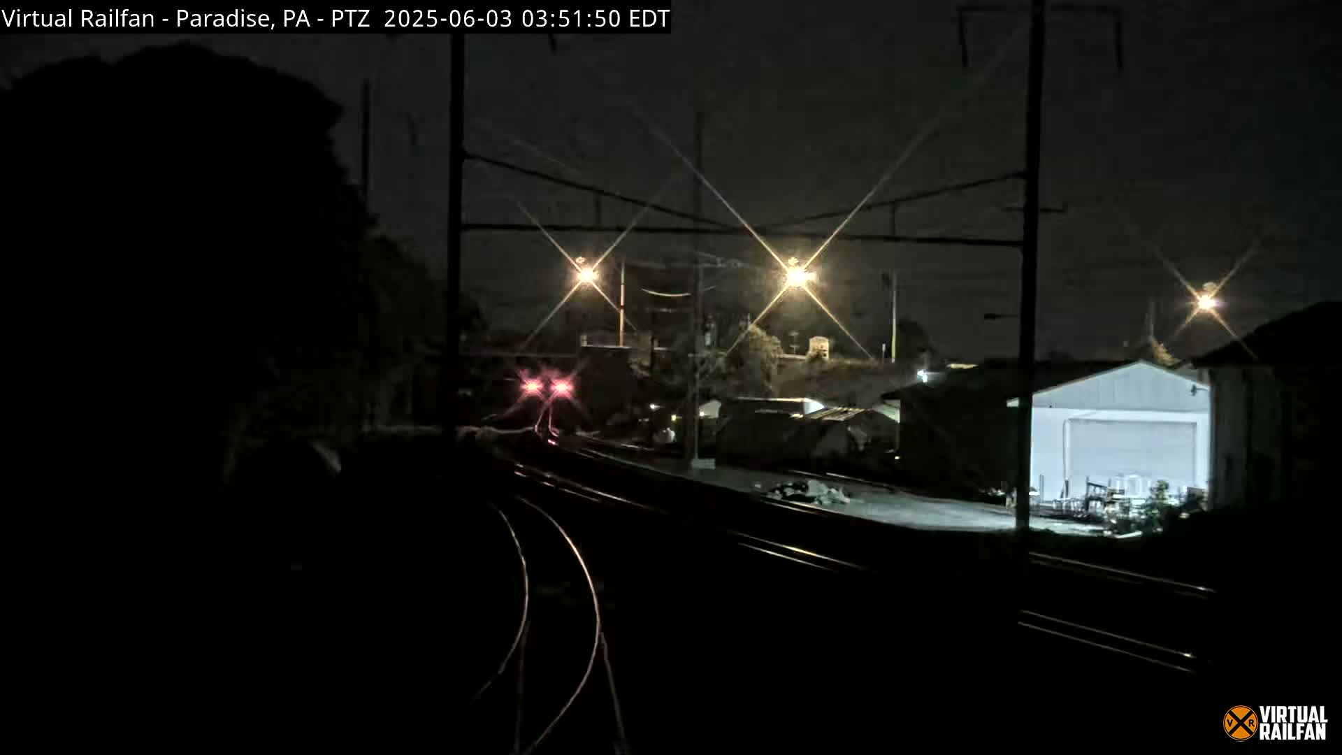 A nighttime view of train tracks and industrial buildings, lit by several bright lights.
