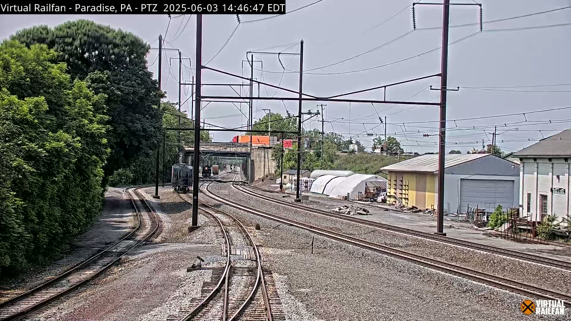 A mostly sunny day shows multiple sets of train tracks converging near a small train, buildings, and lush green trees under a mostly clear sky.