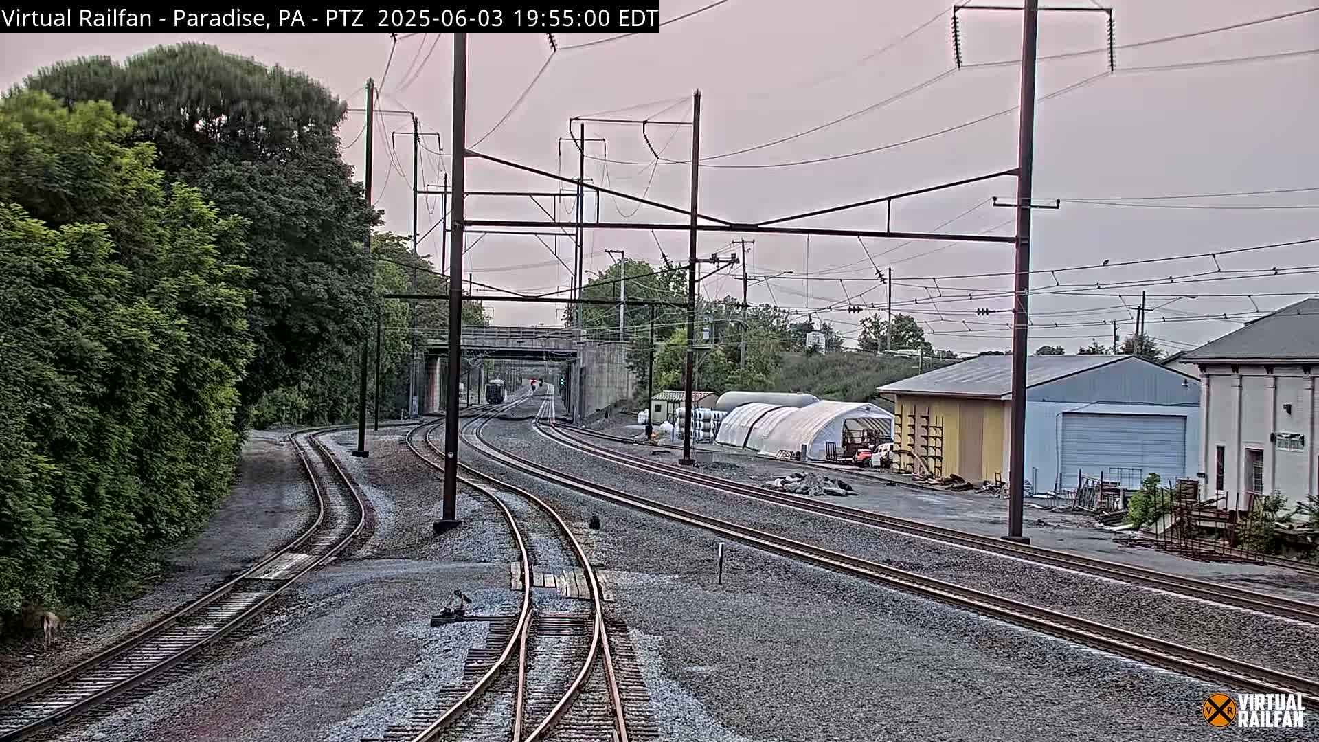 Multiple train tracks extend into the distance under an overcast sky, past a building and a wooded area.