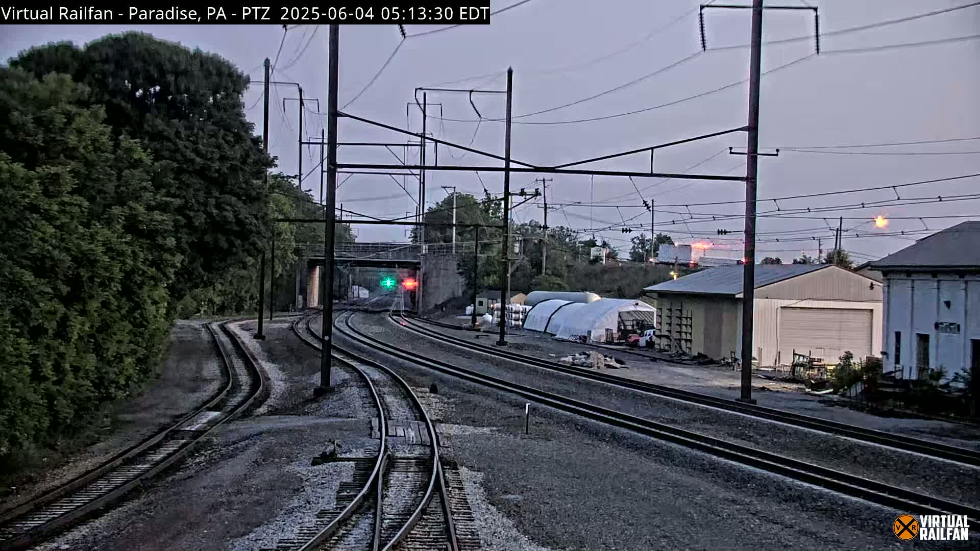 Several sets of train tracks converge near a low building and a large greenhouse under a dusky sky.