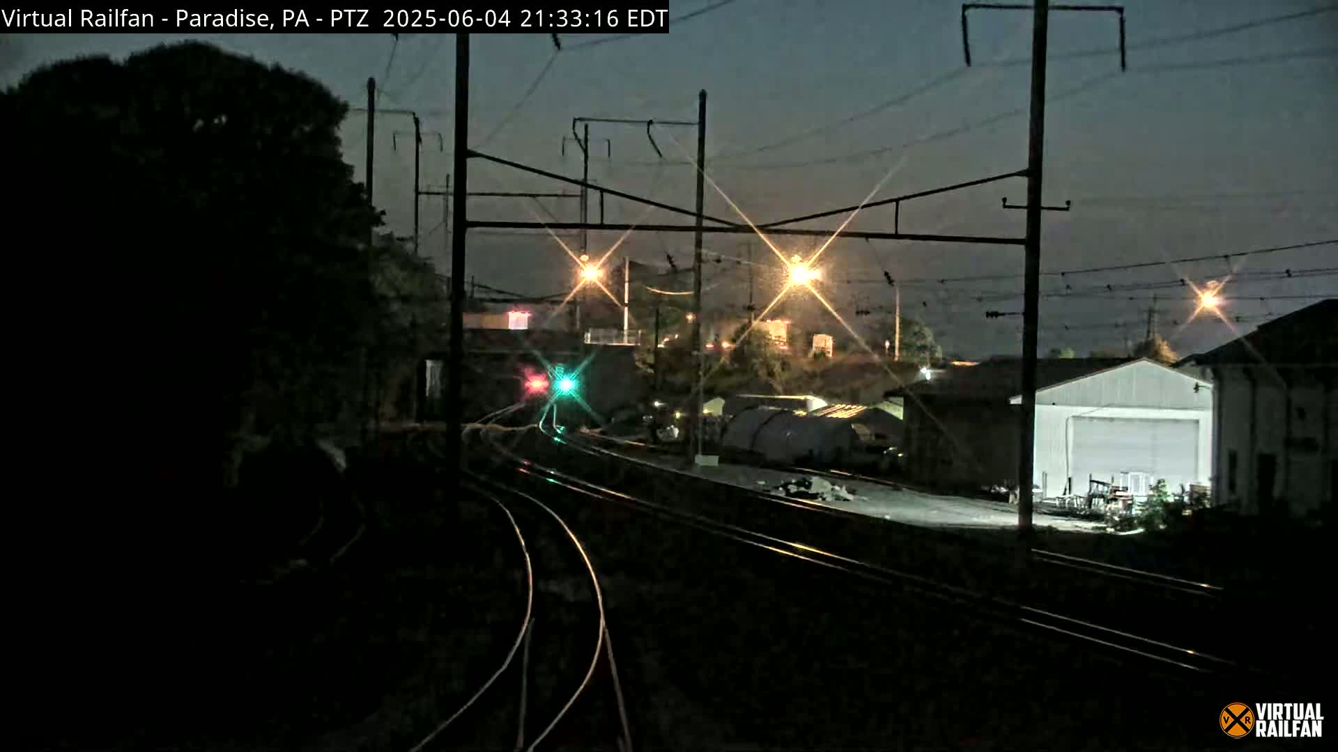 A nighttime view of a railroad yard with converging train tracks, illuminated by several lights, under a dark sky.
